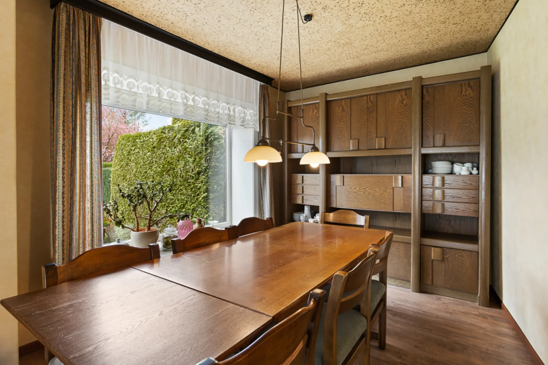 Esszimmer mit großem Holztisch, Stühlen und Holzregal, daneben großes Fenster mit Blick ins Grüne.