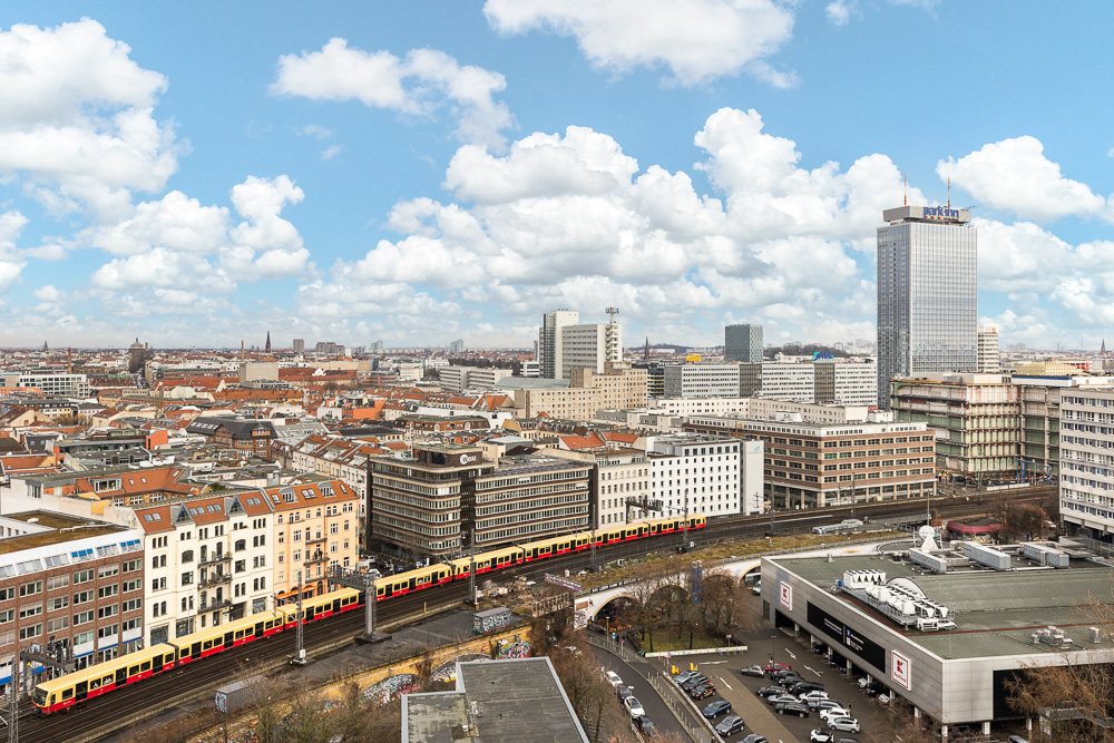 Stadtansicht mit Wohngebäuden, Hochhäusern, S-Bahn und Supermarkt bei blauem Himmel.
