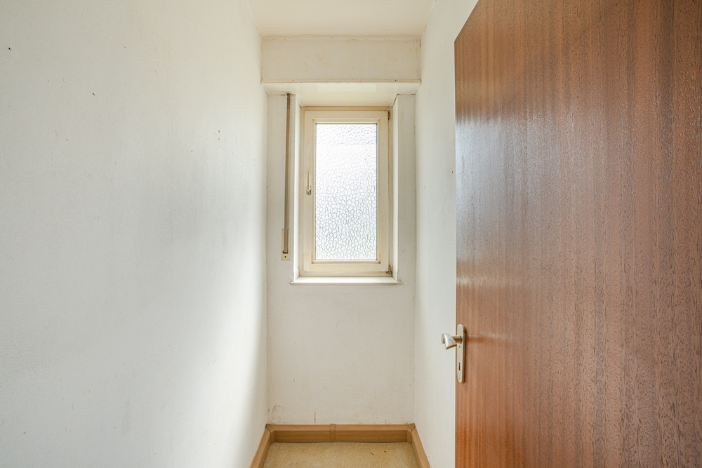 Kleiner, leerer Raum mit weiß gestrichenen Wänden und einem Fenster am Ende.