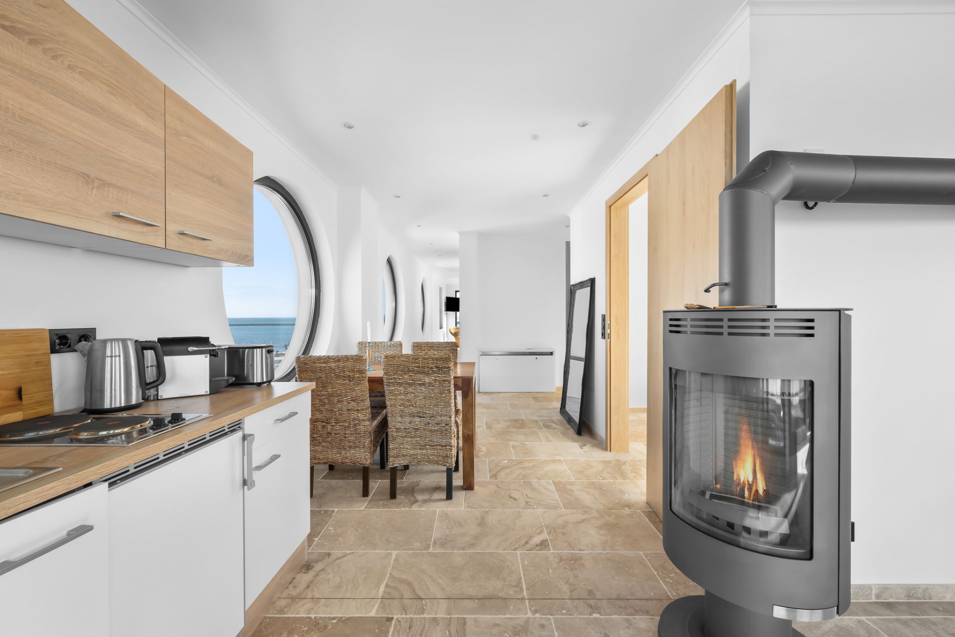 Moderne offene Küche mit Holzofen, Esstisch und runden Fenstern mit Blick aufs Meer.
