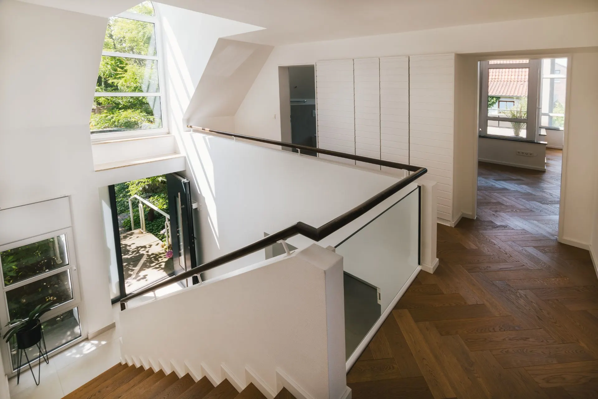 Heller Flur mit Treppe, Glasgeländer und großen Fenstern, Holzboden, Zugang zu weiteren Räumen.