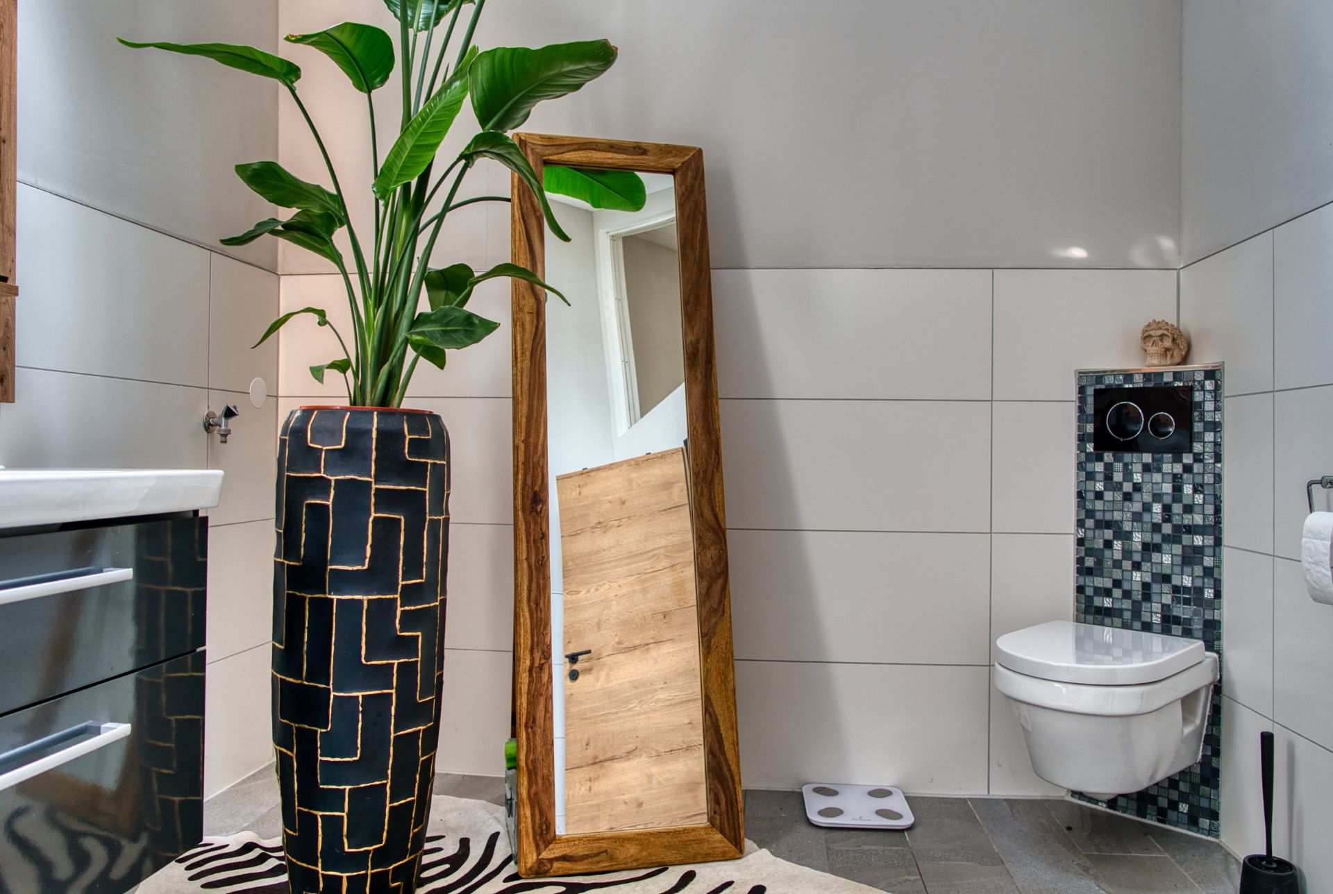 Modernes Badezimmer mit Pflanze im großen Vase, Spiegel und wandhängender Toilette.