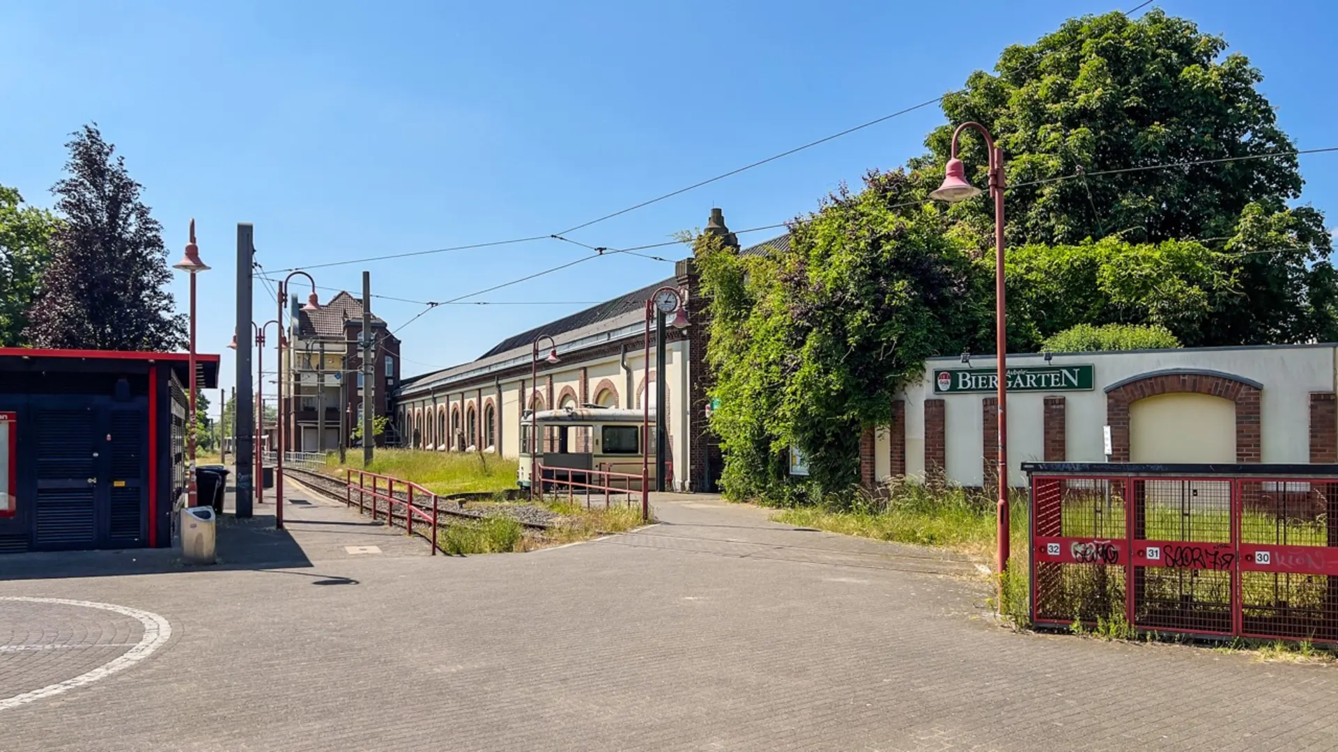 Gepflasterter Platz mit Schienen, altem Gebäude, Bäumen und einem Schild Biergarten.