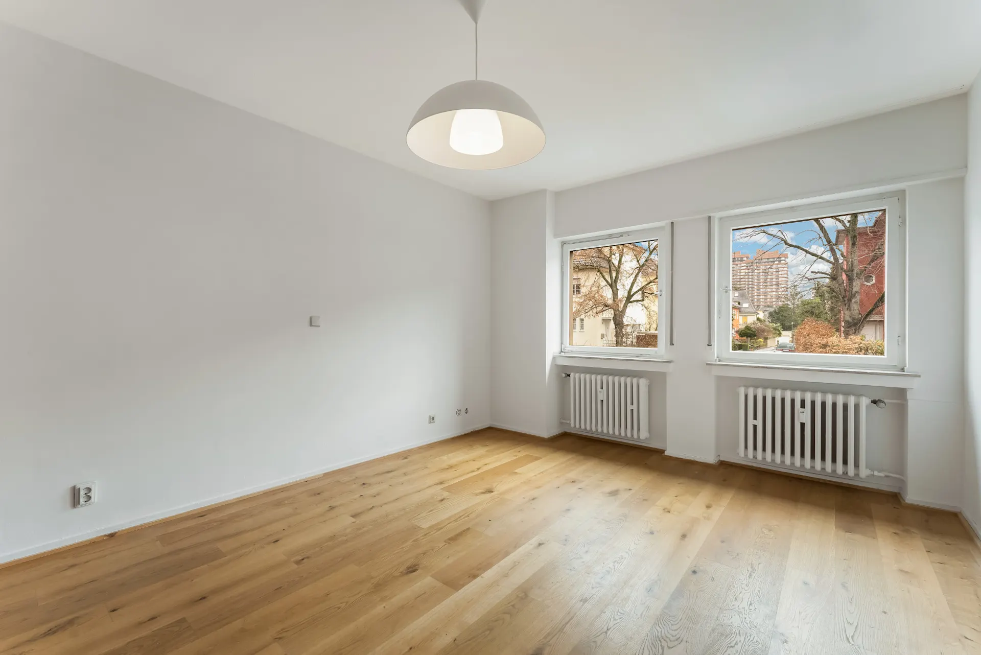 Helles, leeres Zimmer mit Holzfußboden, zwei Fenstern und weißer Deckenlampe.