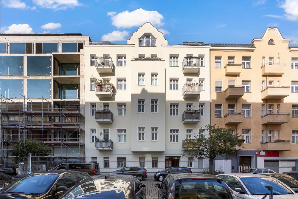 Mehrfamilienhaus mit Balkonen, geparkte Autos davor, links angrenzendes Gebäude im Bau.