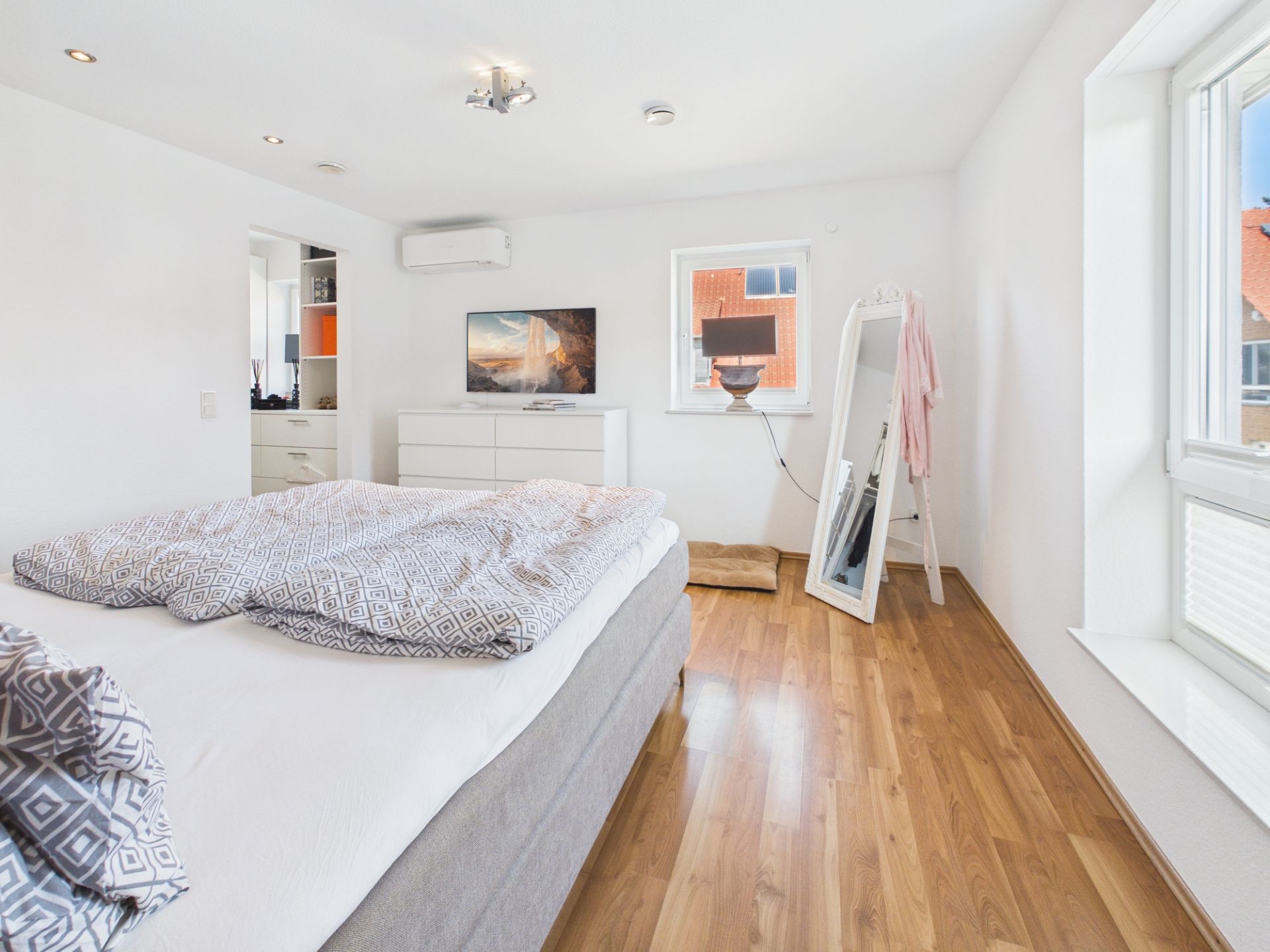 Helles Schlafzimmer mit Doppelbett, Kommode, Spiegel und großen Fenstern.