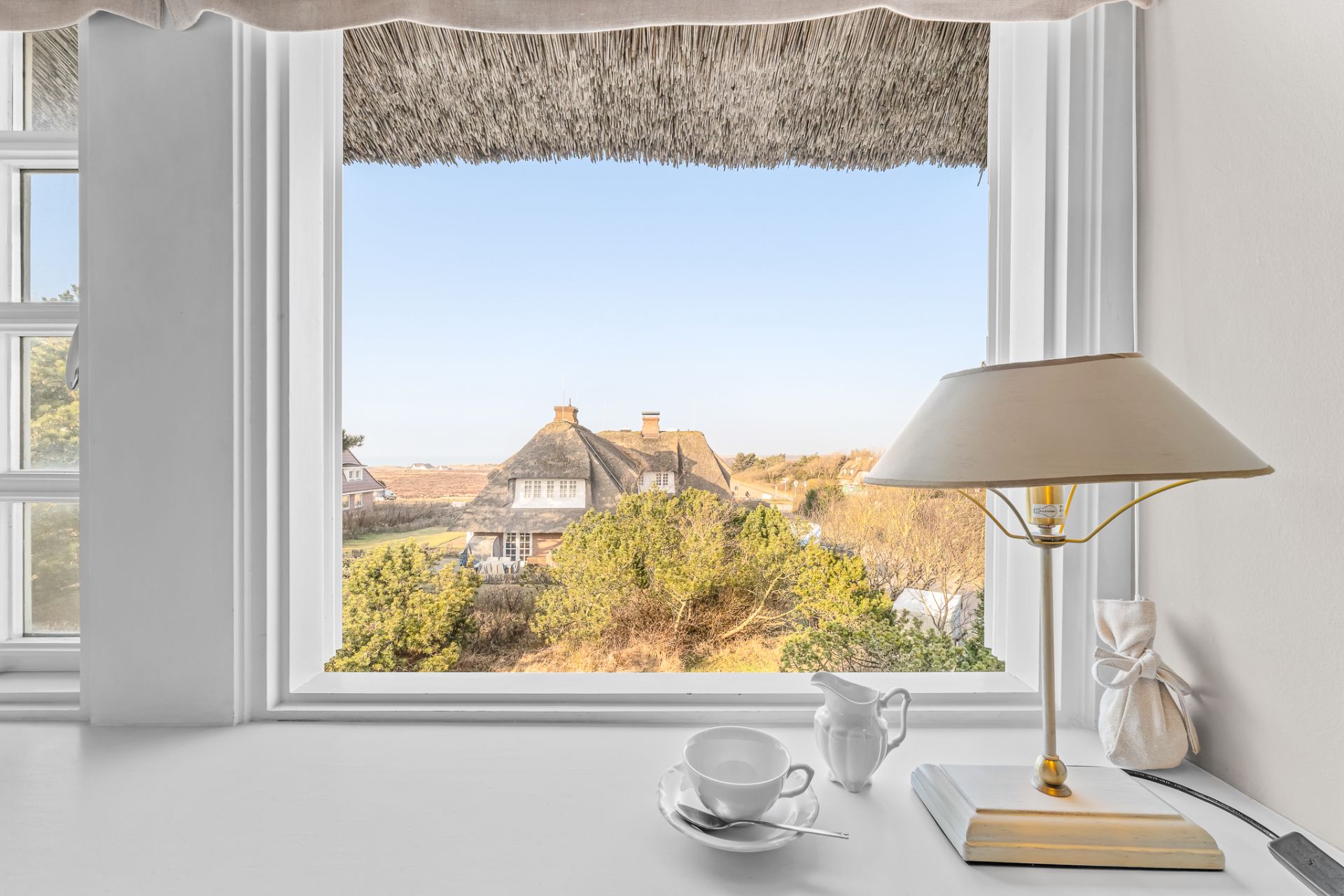 Blick aus Fenster mit Tischlampe und Teetasse auf Haus mit Reetdach und Bäumen.