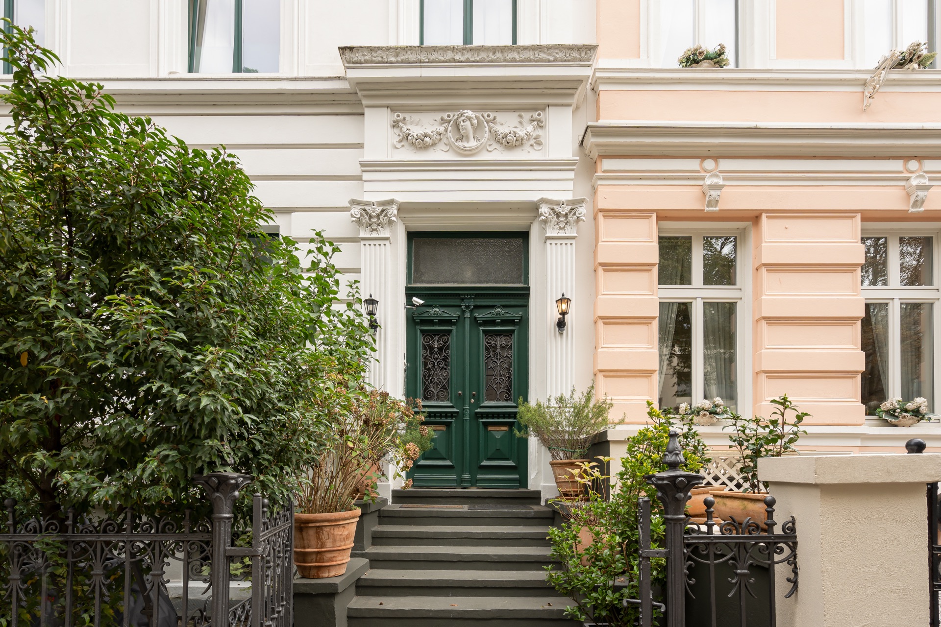 Vorgarten mit grünen Pflanzen und Treppe zu einer verzierten dunklen Haustür.