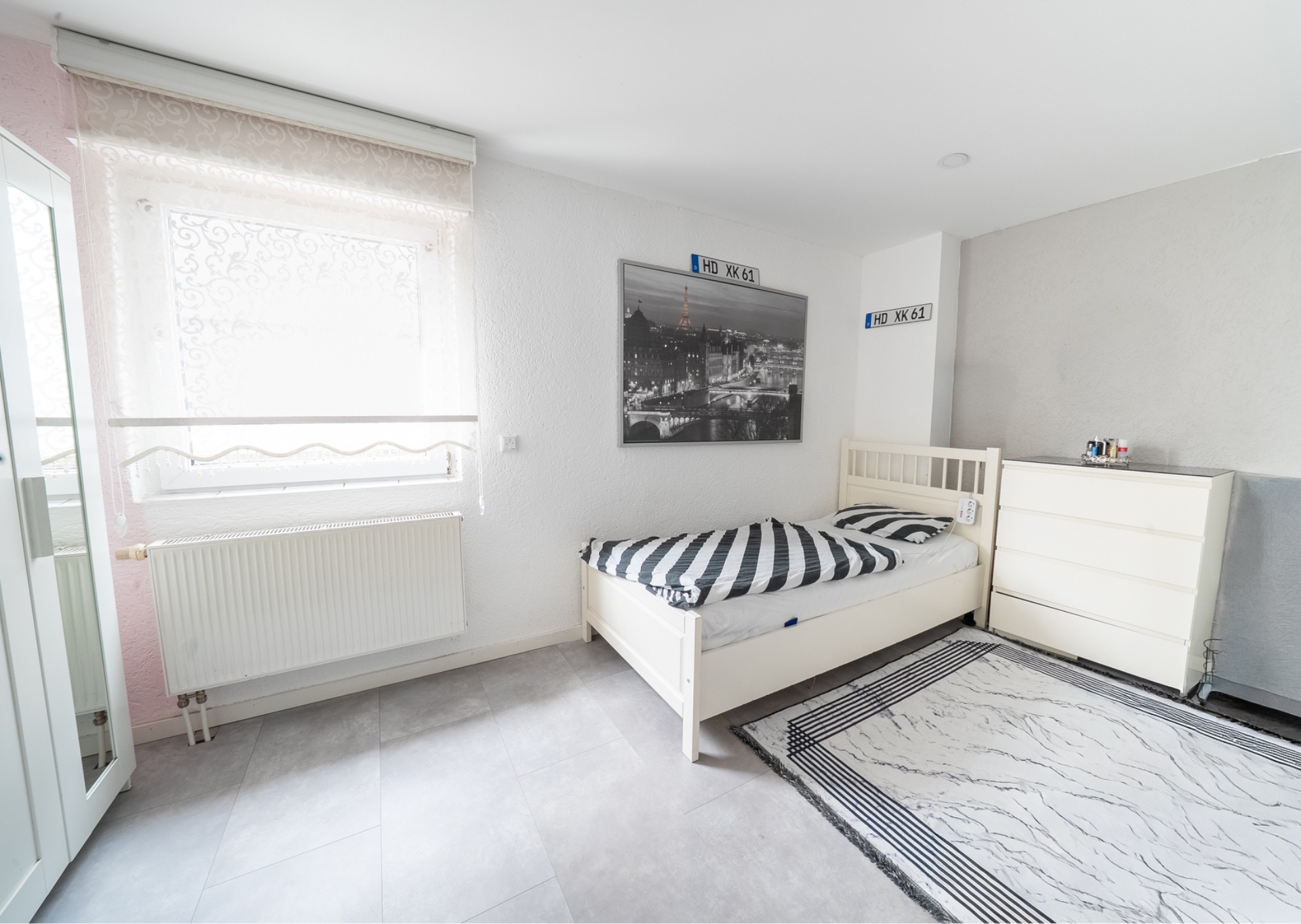 Helles Schlafzimmer mit Einzelbett, Kommode, Fenster und Teppich.
