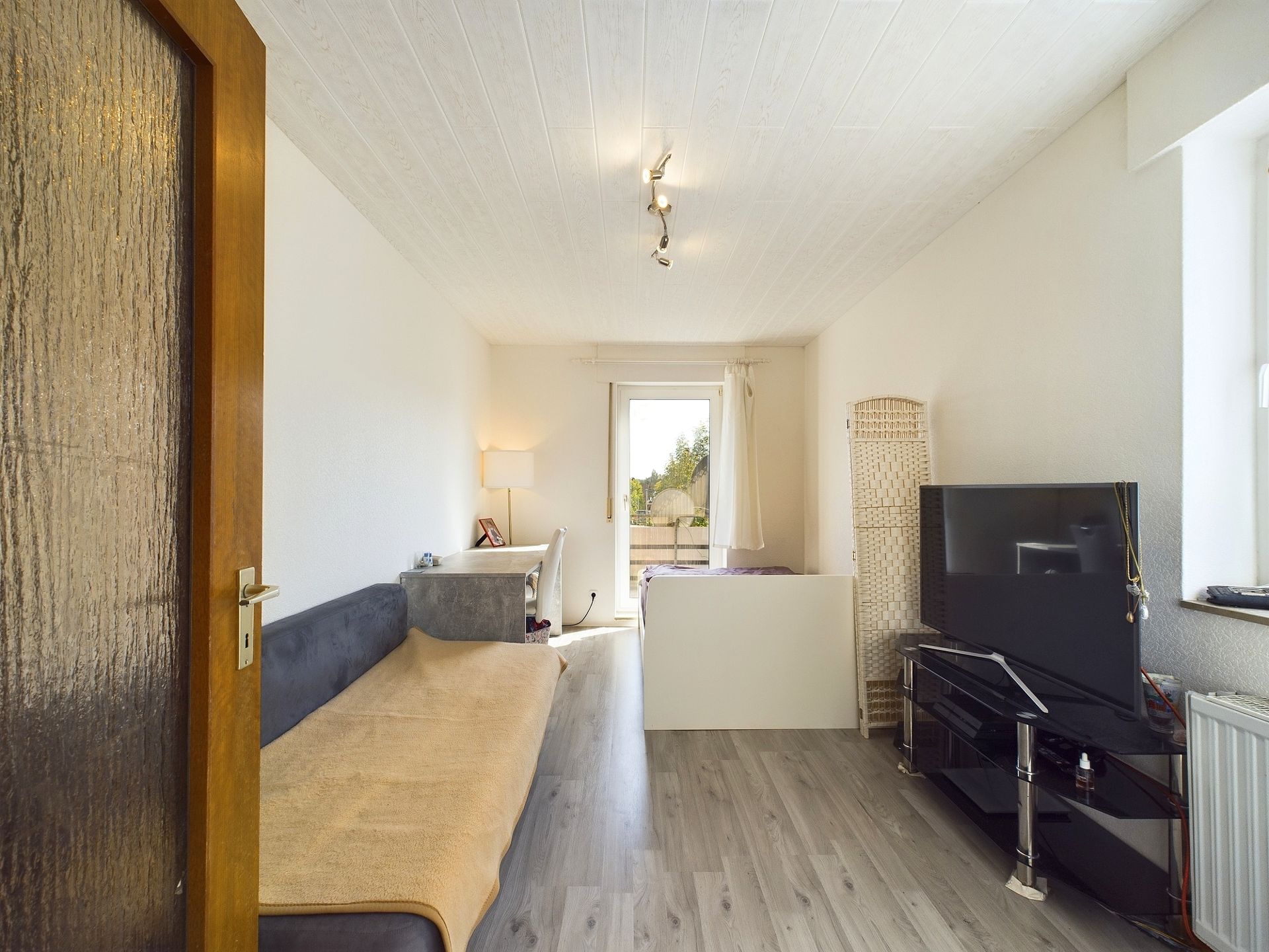Helles Wohnzimmer mit Sofa, Fernseher und Glastür zum Balkon.