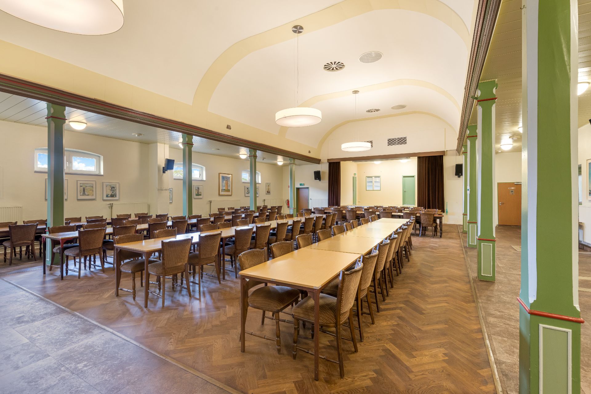 Großer Saal mit vielen Tischen und Stühlen, Holzboden und hellen Wänden.