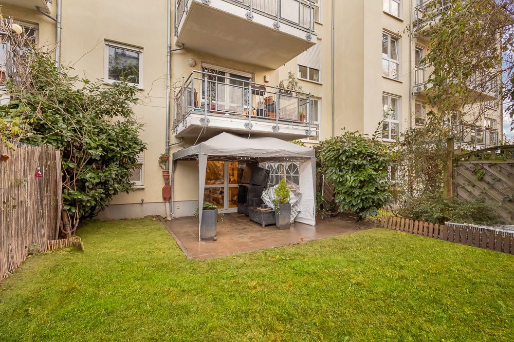 Garten mit Rasen, Pavillon und Sitzmöbeln vor einem Wohngebäude mit Balkonen.