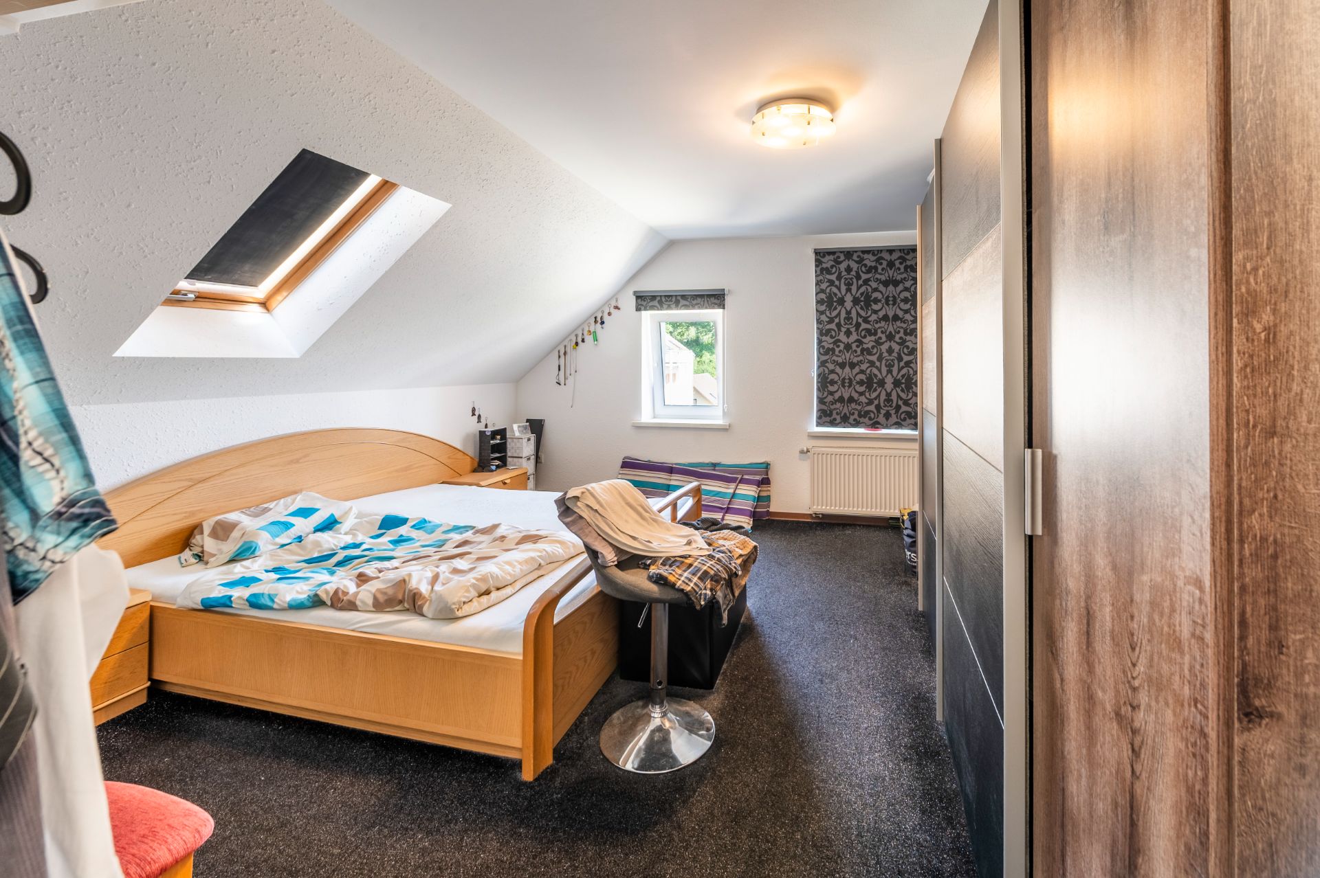 Helles Schlafzimmer mit Dachfenster, Holzbett, Schrank und Sitzgelegenheit.