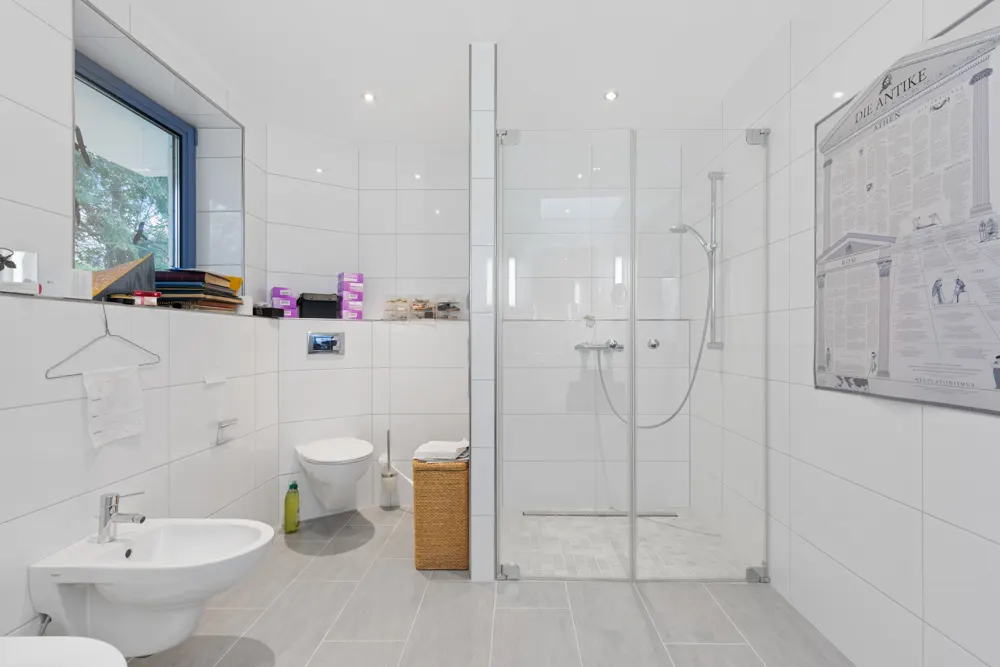 Modernes Badezimmer mit Dusche, WC, Bidet und Fenster, helle Fliesen und wandhängendem Regal.