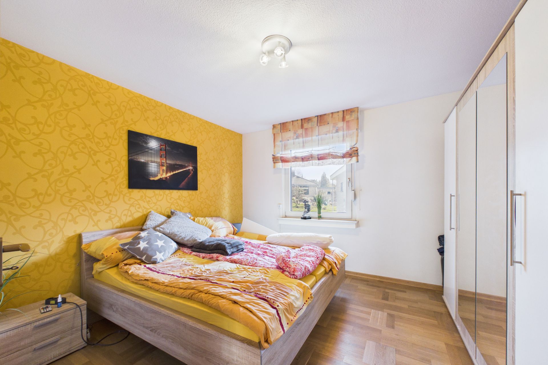 Helles Schlafzimmer mit gelber Akzentwand, Doppelbett, Fenster und großem Kleiderschrank mit Spiegel.