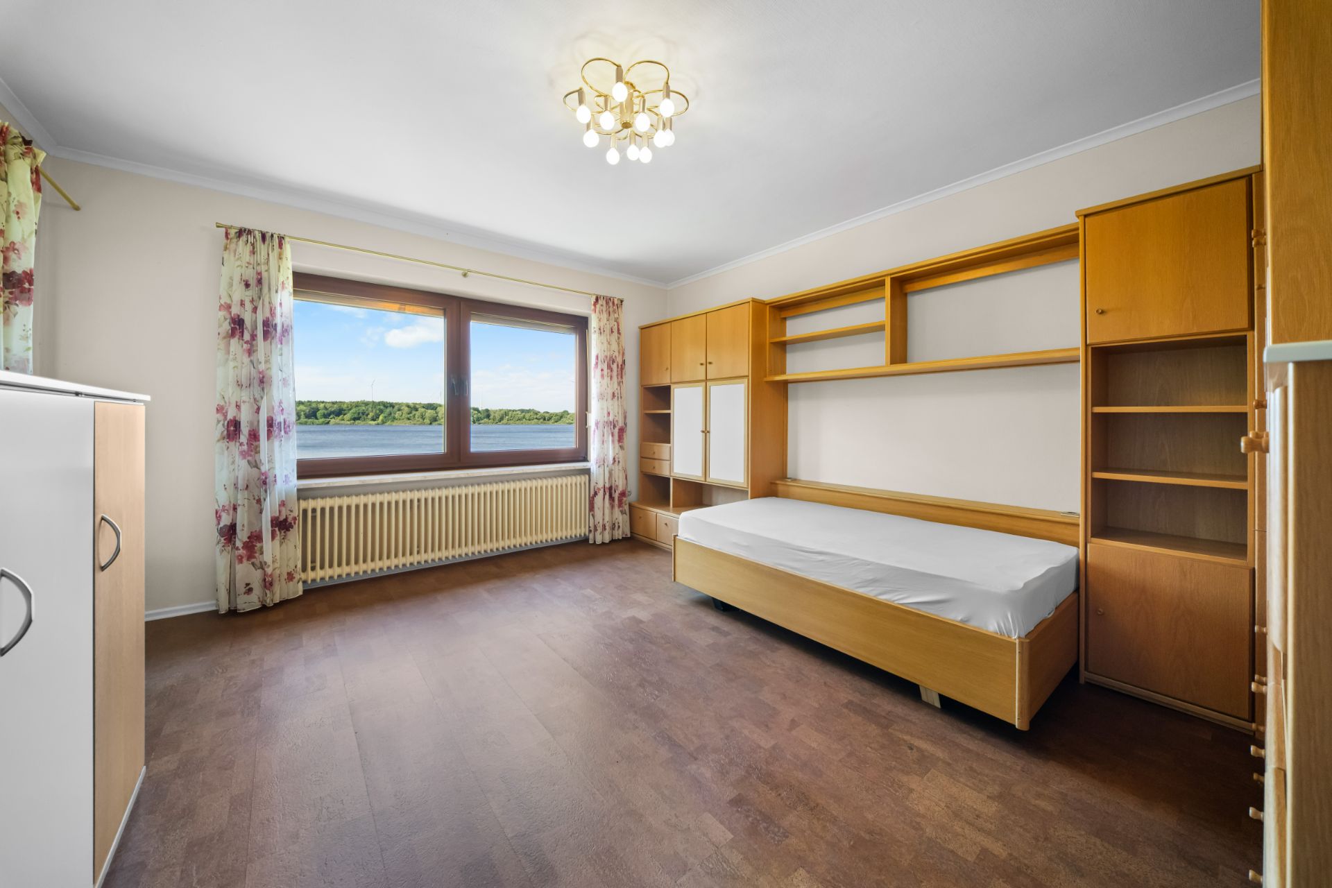 Helles Schlafzimmer mit Einzelbett, Einbauschrank, Regalwand und großem Fenster mit Blick ins Grüne.