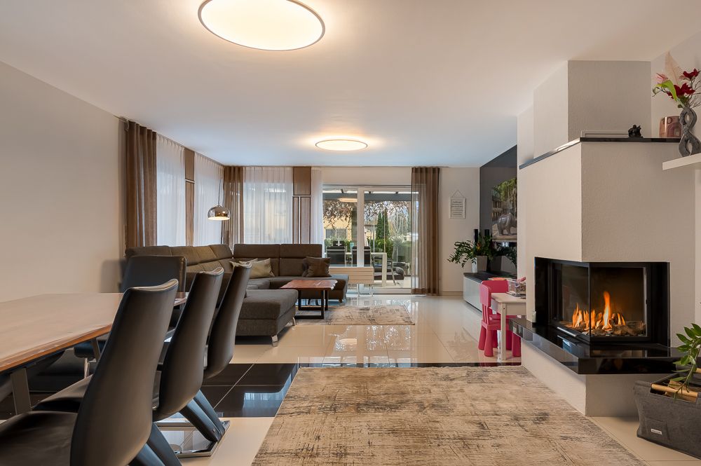 Modernes Wohnzimmer mit Kamin, Ecksofa, Esstisch und großen Fenstern.