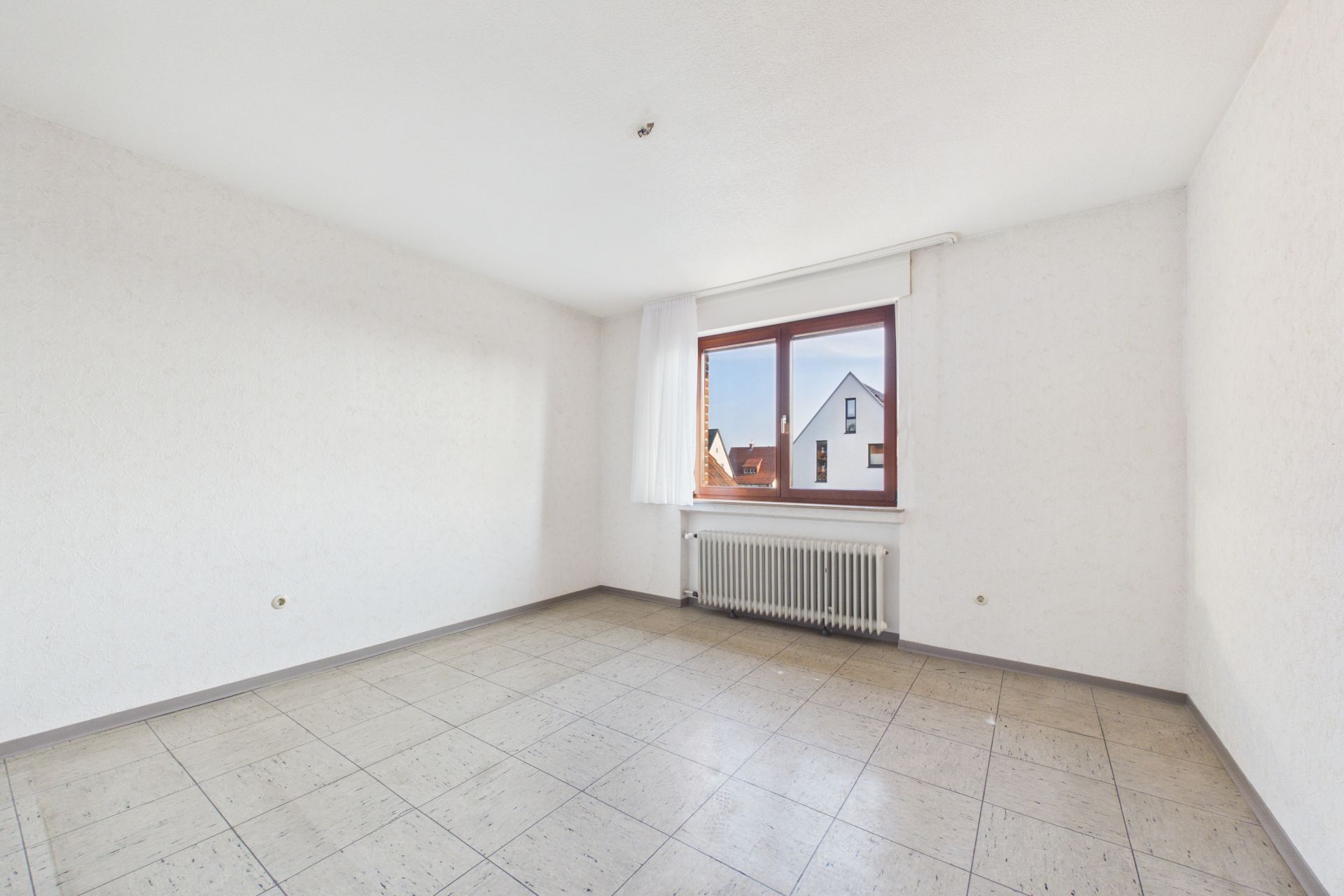 Leeres Zimmer mit Fliesenboden, weißer Wand und großem Fenster mit Blick nach draußen.