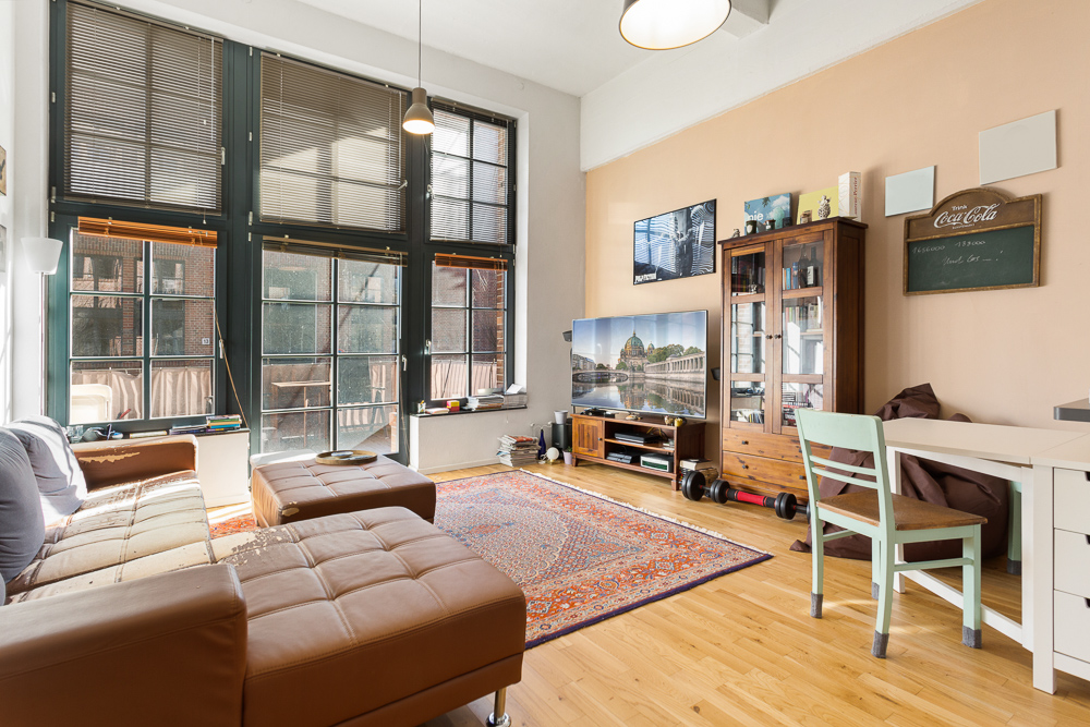 Wohnzimmer mit großen Fenstern, braunem Sofa, Teppich, TV, Holzschrank und hellem Holzboden.