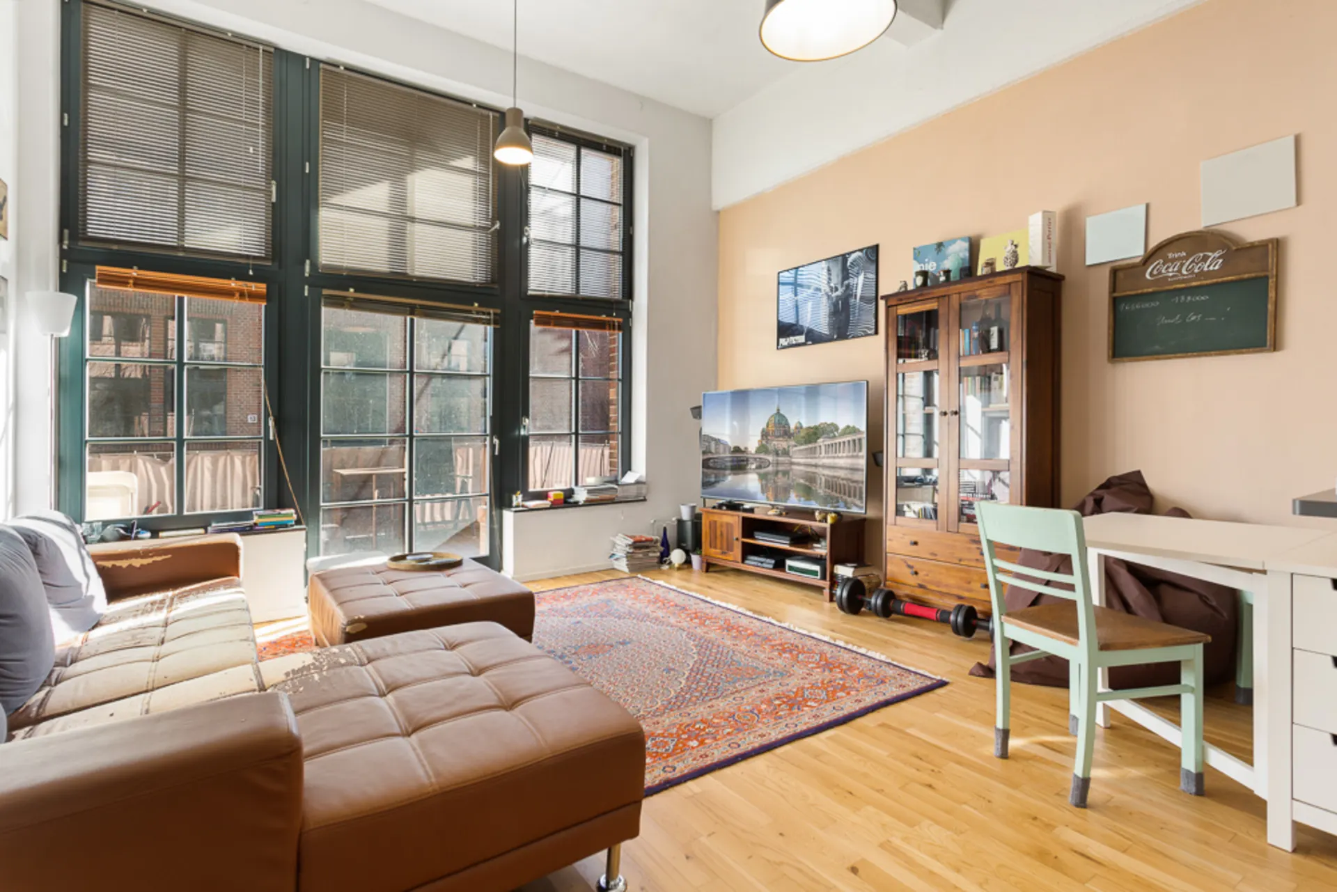 Wohnzimmer mit großen Fenstern, braunem Sofa, Teppich, TV, Holzschrank und hellem Holzboden.