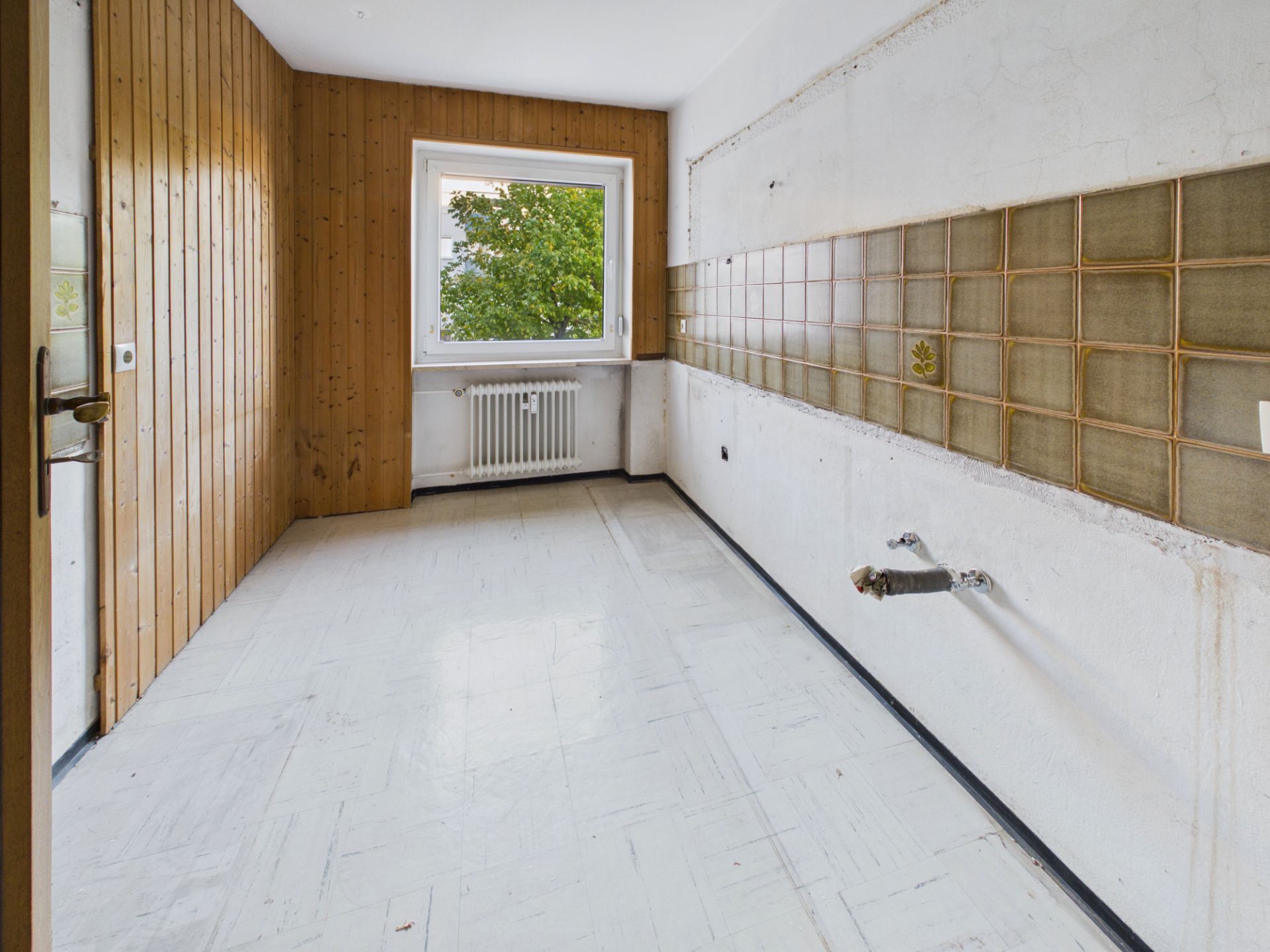 Leerer Raum mit Fliesen an einer Wand, Holzverkleidung und Fenster mit Blick ins Grüne.