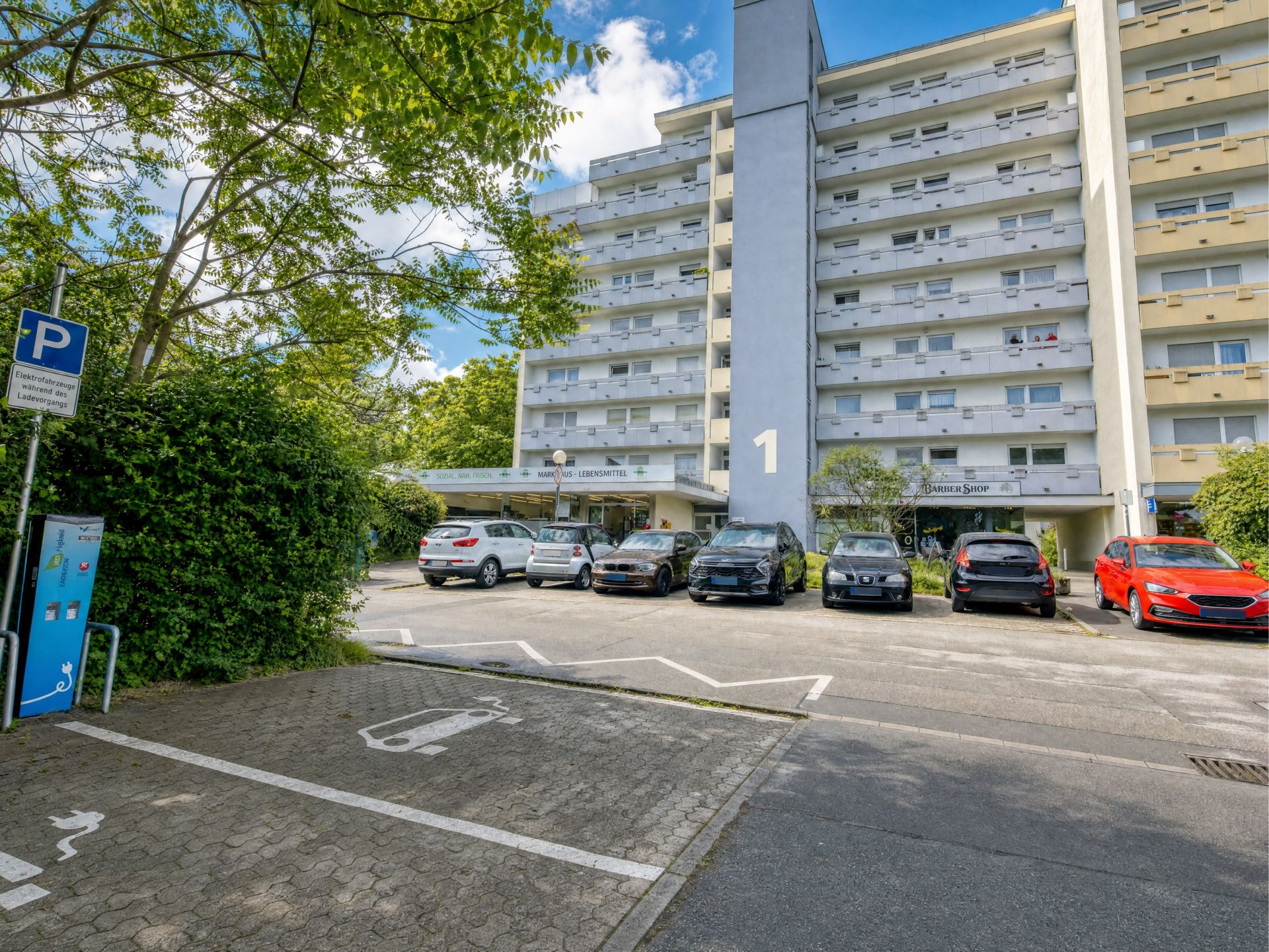 Mehrstöckiges Wohngebäude mit Parkplätzen und Autos davor, Rollstuhlparkplatz im Vordergrund.
