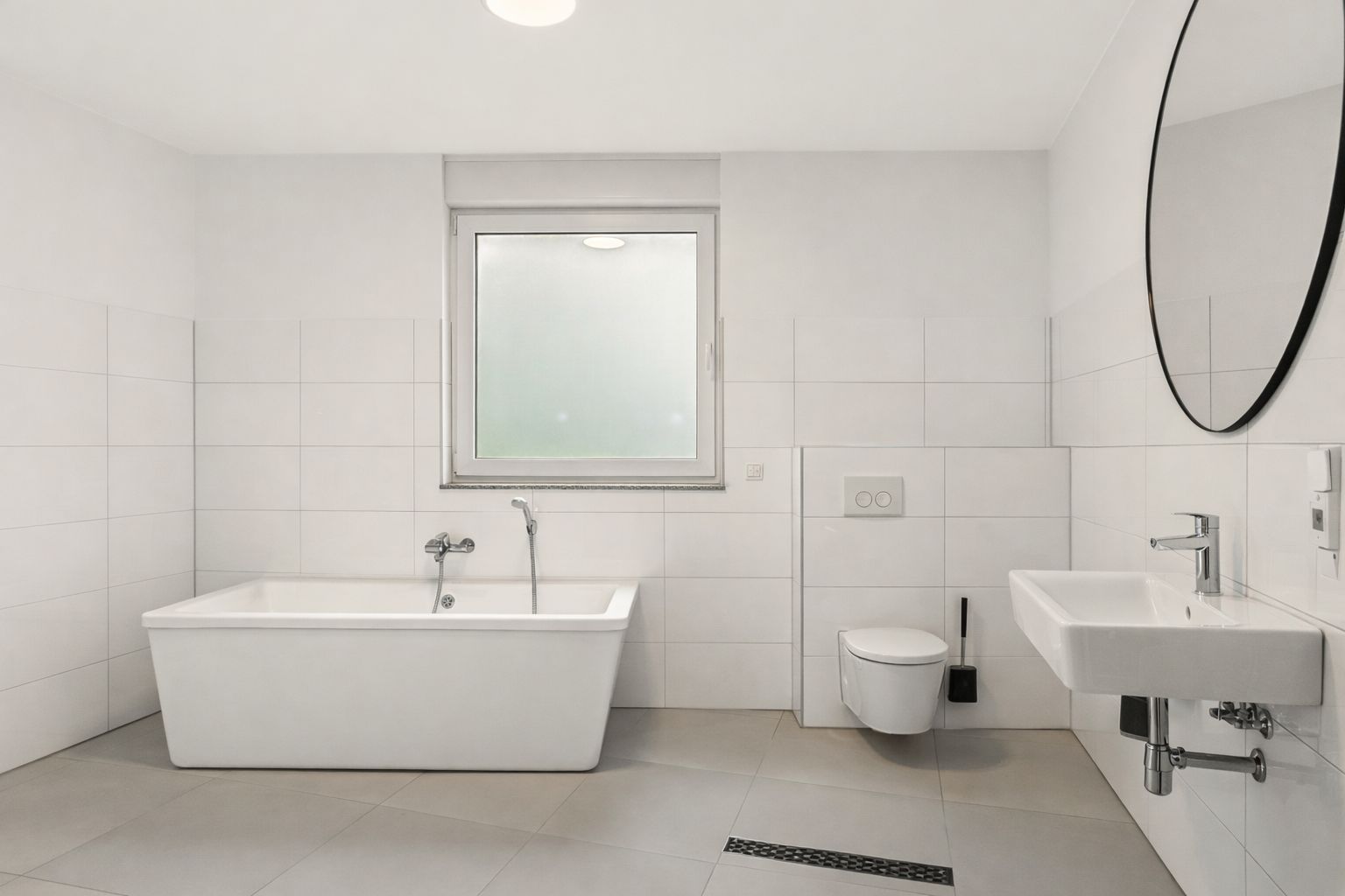 Modernes Badezimmer mit Badewanne, Fenster, Wandtoilette, Waschbecken und rundem Spiegel.