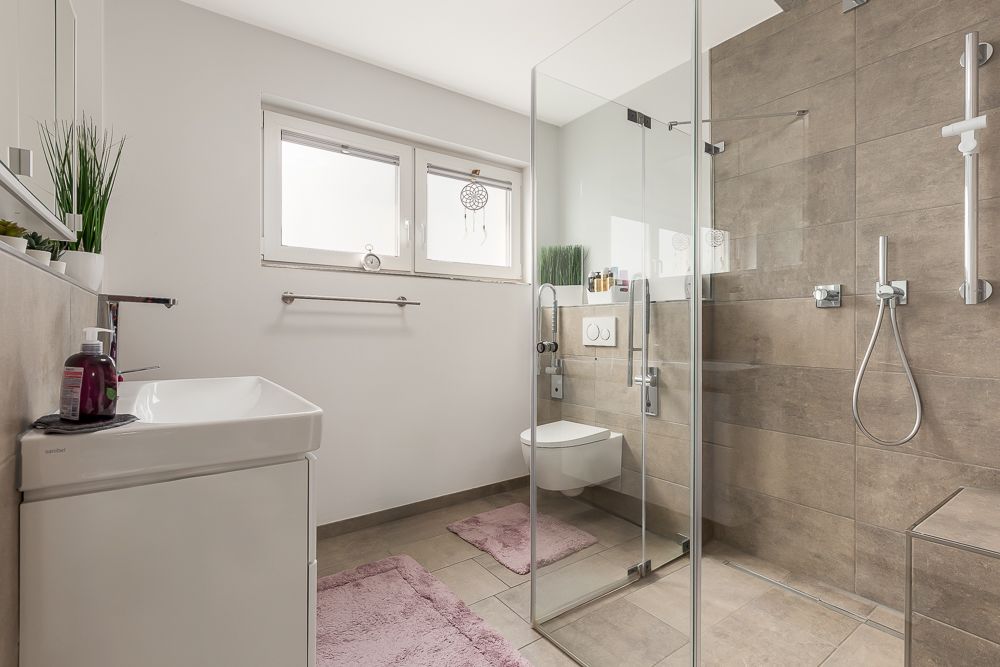 Modernes Bad mit Glasdusche, wandhängender Toilette und zwei Fenstern, rosa Teppichen auf dem Boden.