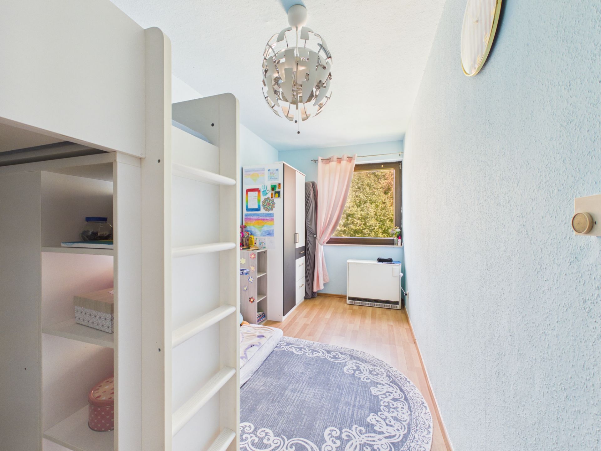 Helles Kinderzimmer mit Hochbett, Schrank, Teppich und Fenster mit rosa Vorhängen.