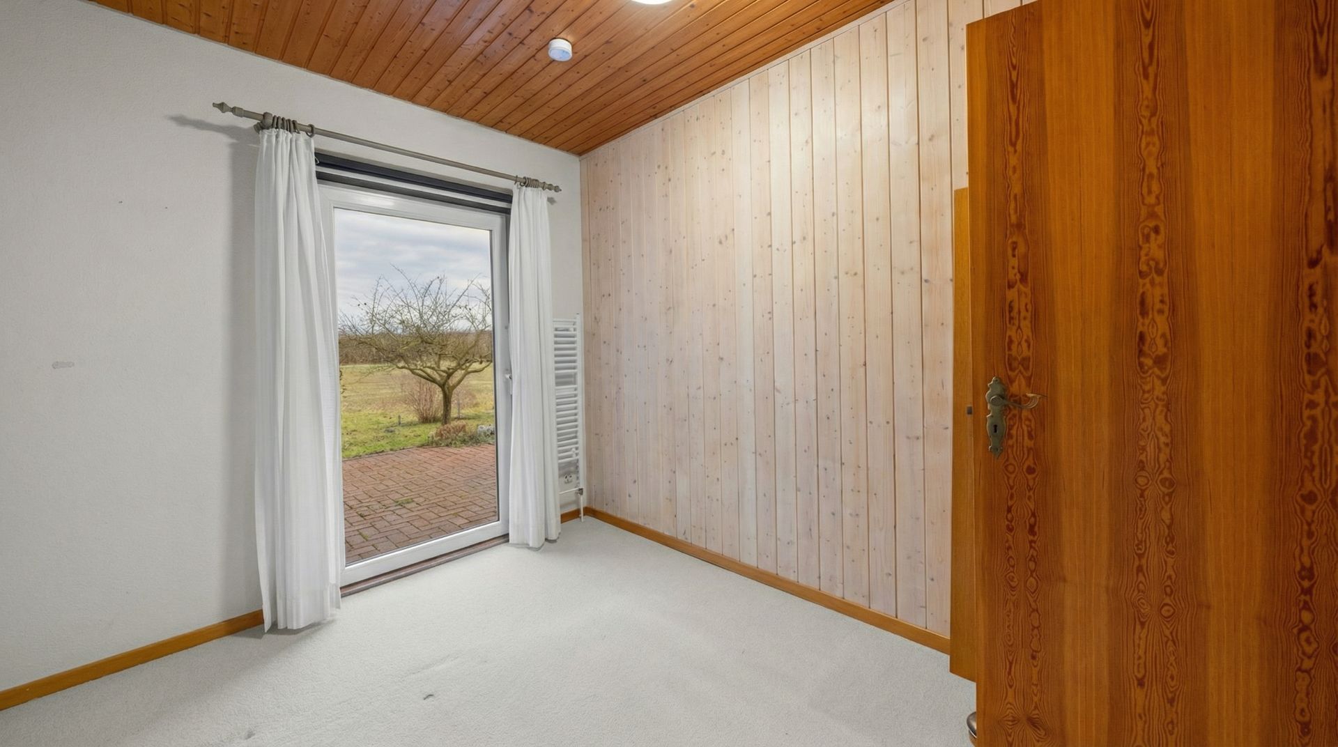 Heller Raum mit Holzwand, Holzdecke, Teppichboden und Tür zur Terrasse mit Blick nach draußen.