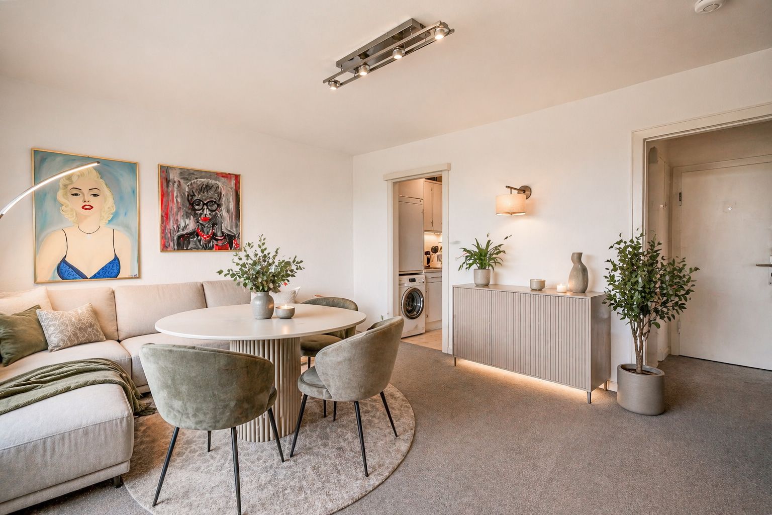 Helles Wohnzimmer mit rundem Esstisch, grünen Stühlen, Sofa, Pflanzen und zwei Bildern an der Wand.