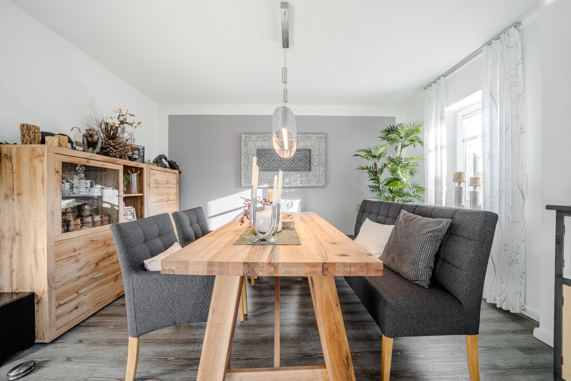 Helles Esszimmer mit Holztisch, grauen Sitzbänken, Vitrine und großem Fenster.