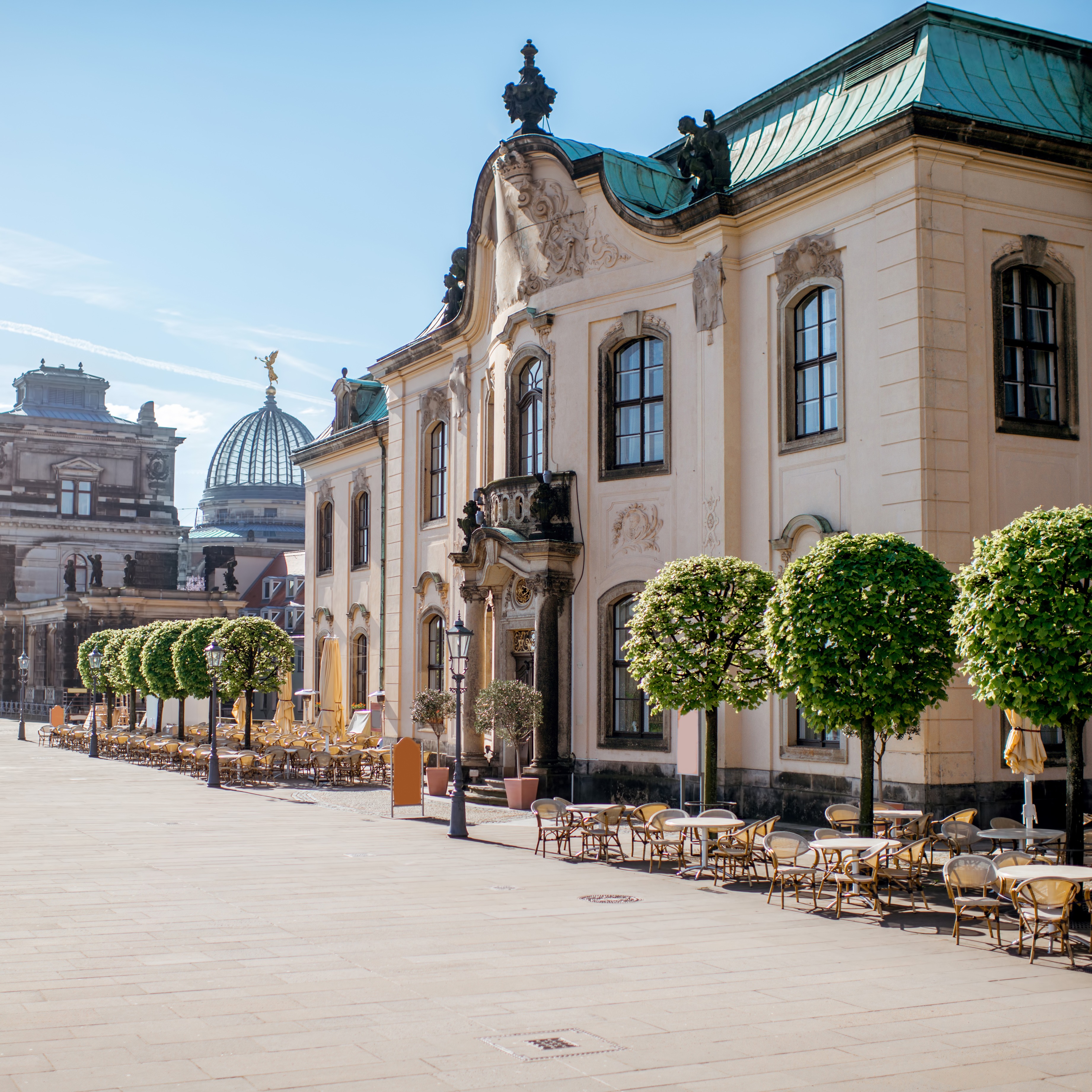 Historisches Gebäude mit Terrasse und Sitzplätzen unter Bäumen an einer sonnigen, gepflasterten Straße.