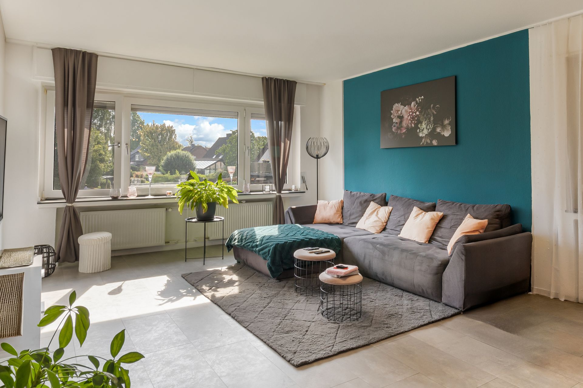 Helles Wohnzimmer mit großem Fenster, grauem Sofa, Teppich und Pflanzen.