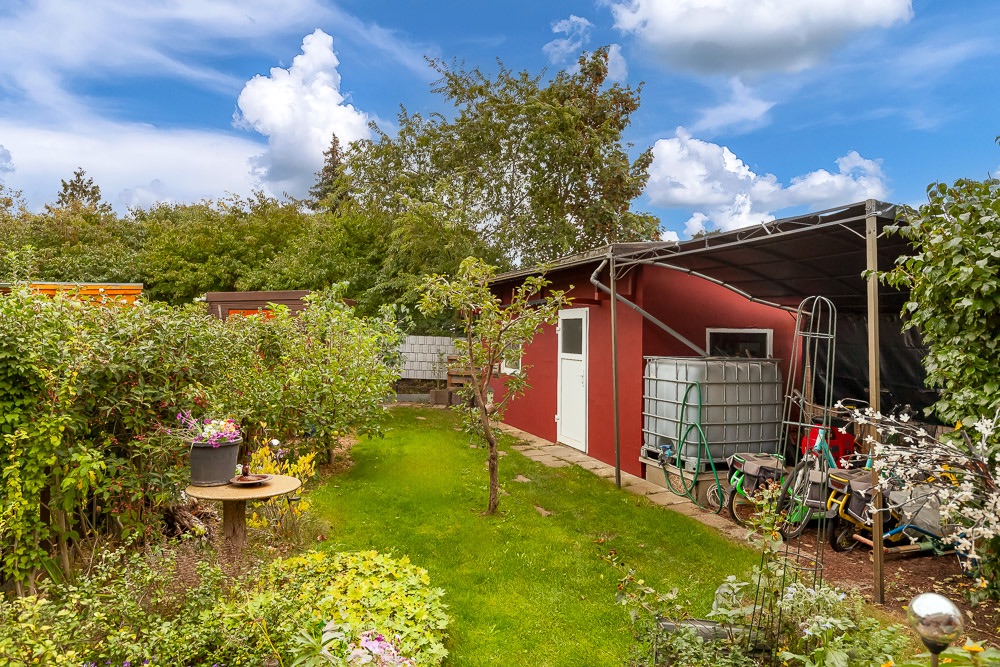 Garten mit Rasen, Bäumen, rotem Nebengebäude und Abstellfläche für Gartengeräte.