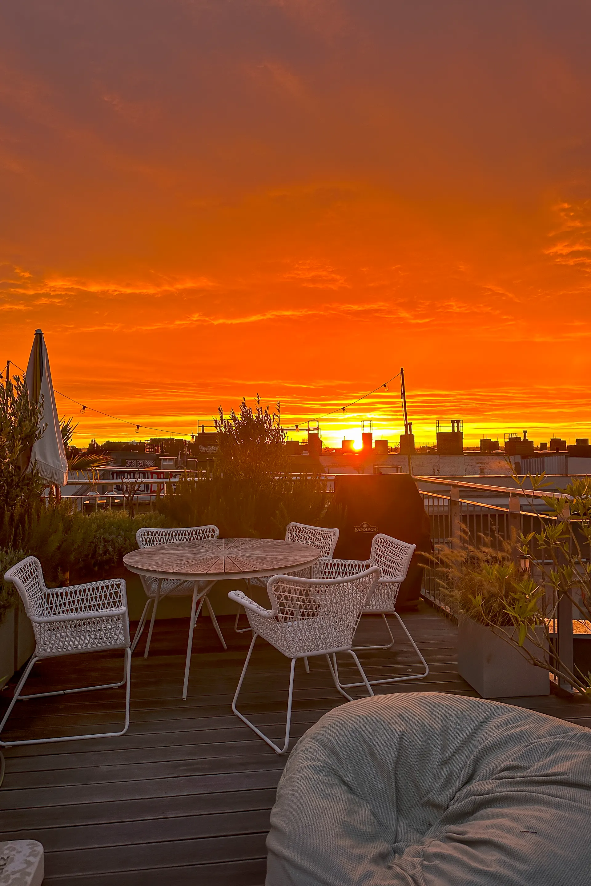 Dachterrasse mit weißen Stühlen, Tisch, Pflanzen und Sonnenuntergang im Hintergrund.