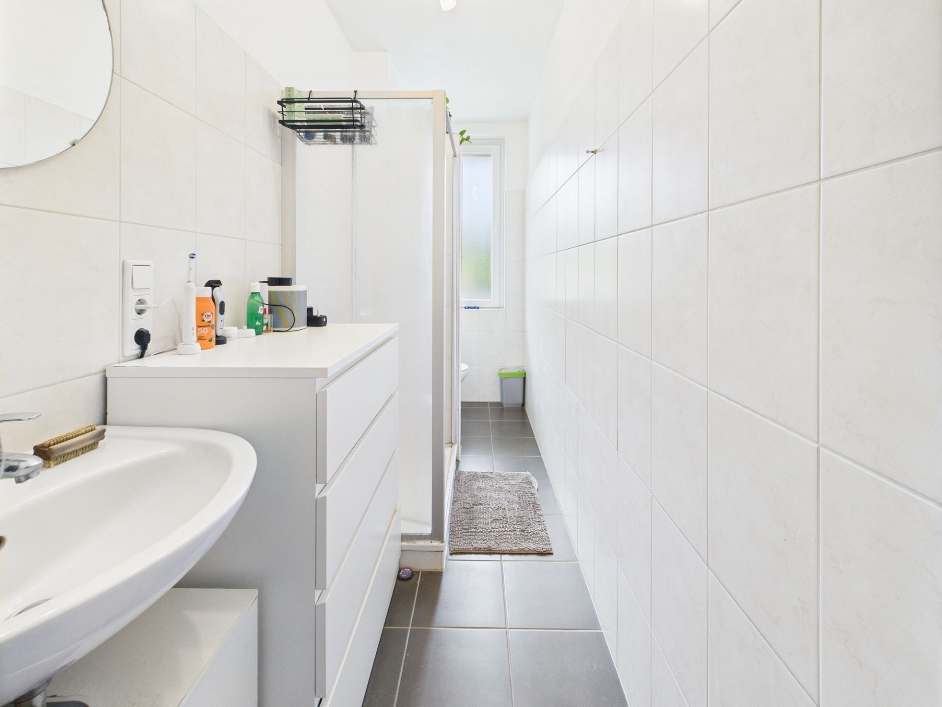 Helles, schmales Badezimmer mit Waschbecken, Kommode, Dusche und Fenster am Ende.