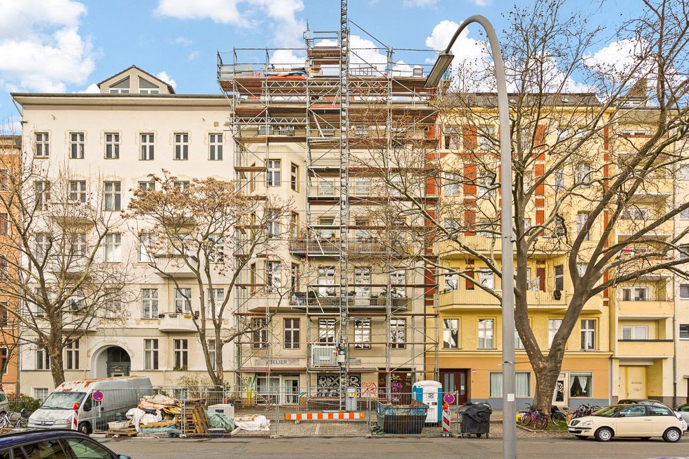Mehrgeschossiges Wohnhaus mit Fassadengerüst und Baustelle davor, Bäume und parkende Autos an der Straße.