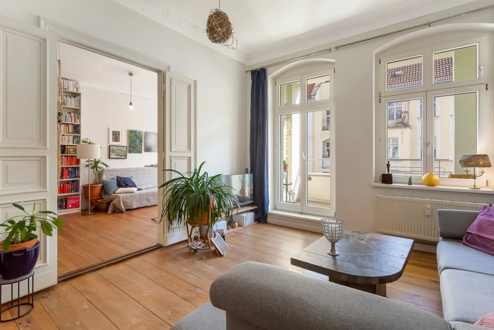 Helles Wohnzimmer mit großen Fenstern, Holzboden, Pflanzen und angrenzendem Raum mit Sofa und Bücherregal.