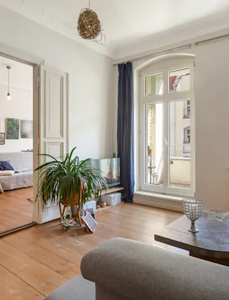 Helles Wohnzimmer mit großen Fenstern, Holzboden, Pflanzen und angrenzendem Raum mit Sofa und Bücherregal.