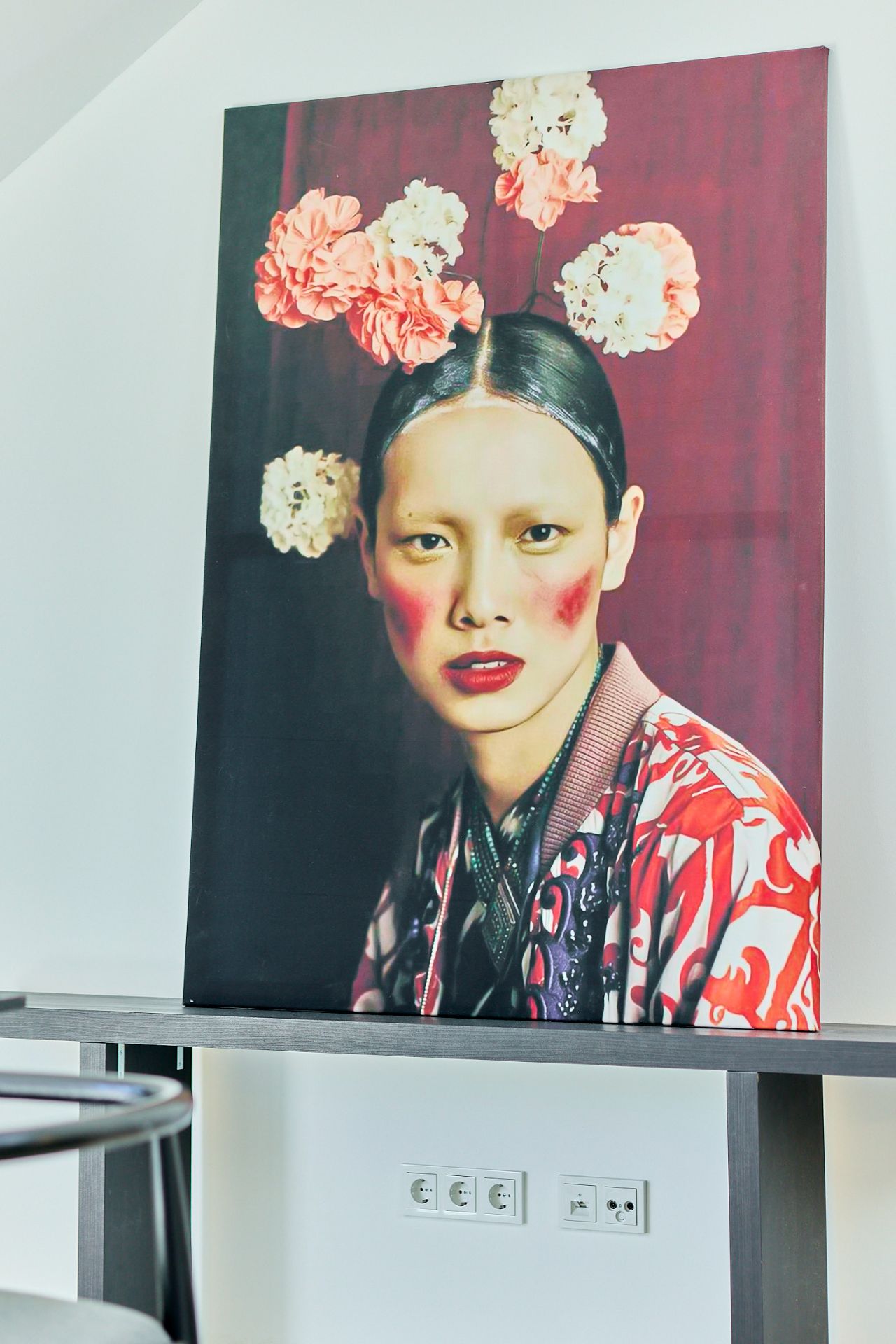 Großes Porträt einer Frau mit Blumen im Haar, auf einem Tisch an einer weißen Wand stehend.