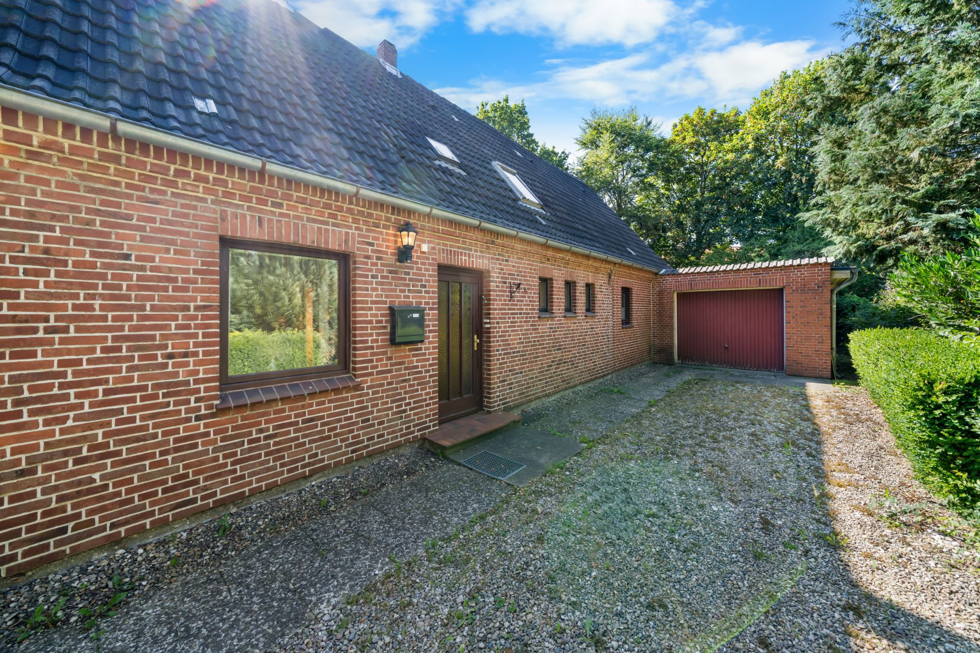 Backsteinhaus mit dunklem Dach, Einfahrt und Garage, umgeben von Bäumen.