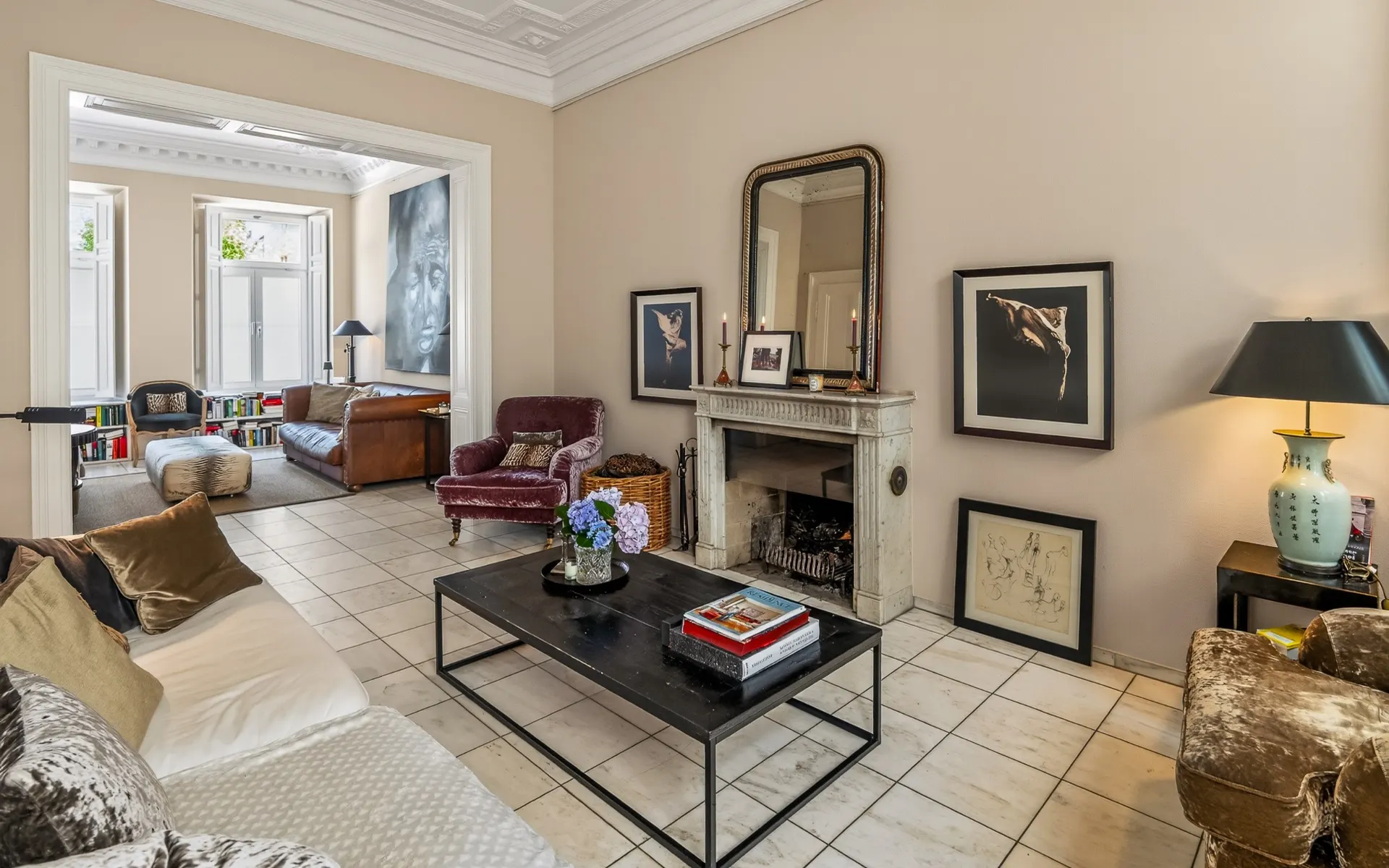 Helles Wohnzimmer mit Kamin, Sofas, Couchtisch und Wandbildern, angrenzendes Zimmer im Hintergrund.