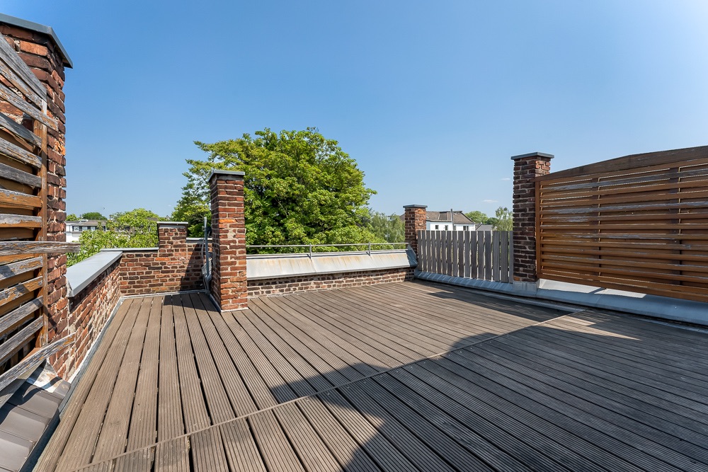 Große Dachterrasse mit Holzboden, Backsteinmauern und Sicht auf Bäume unter blauem Himmel.