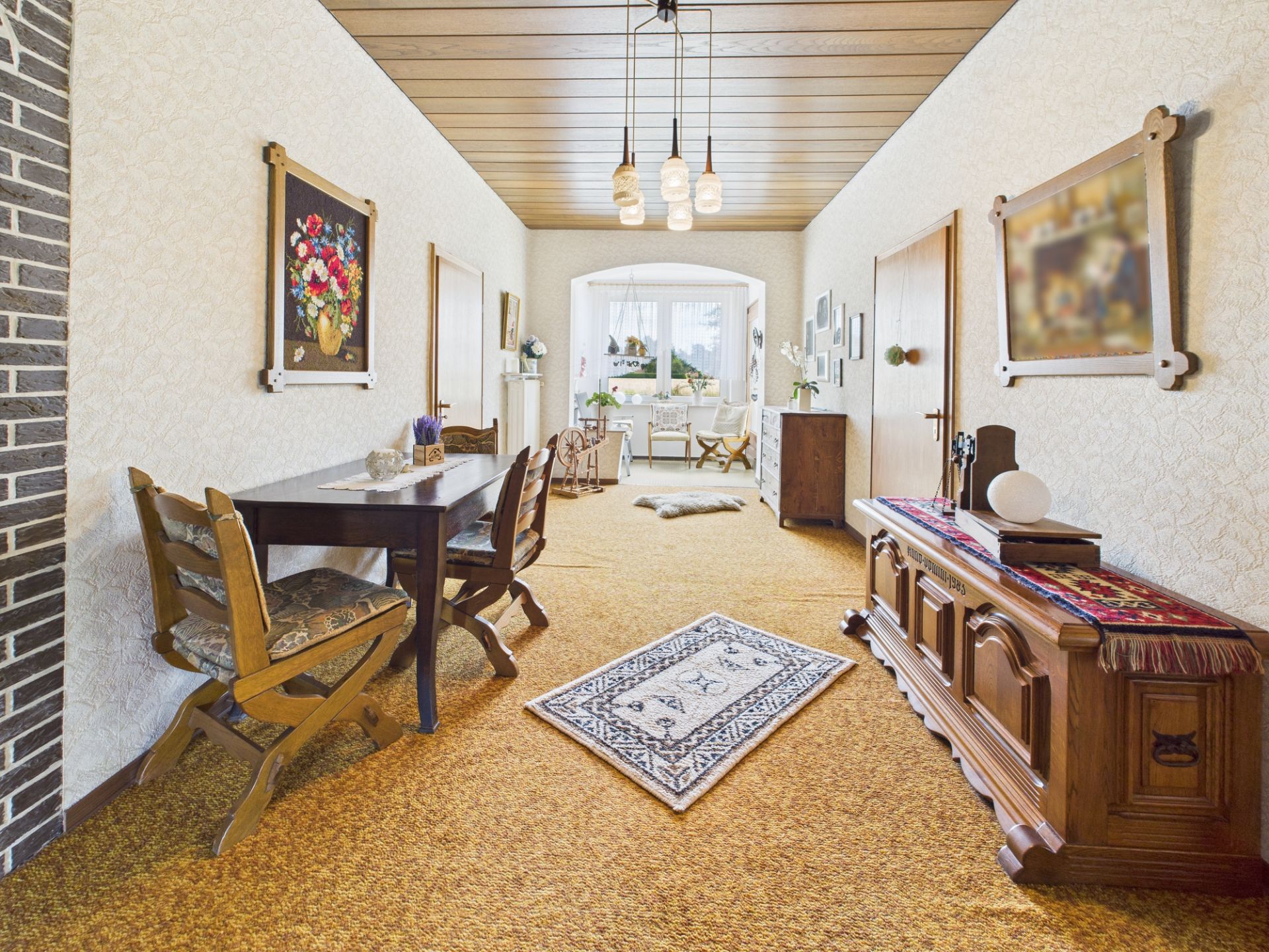 Wohnzimmer mit Holzmöbeln, Teppichboden, Esstisch, Sideboard und Gemälden an den Wänden.