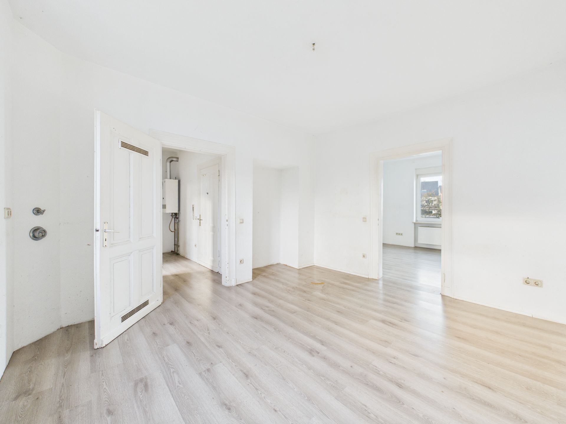 Helles, leeres Zimmer mit hellem Laminatboden, weißen Wänden und zwei offenen Türen.