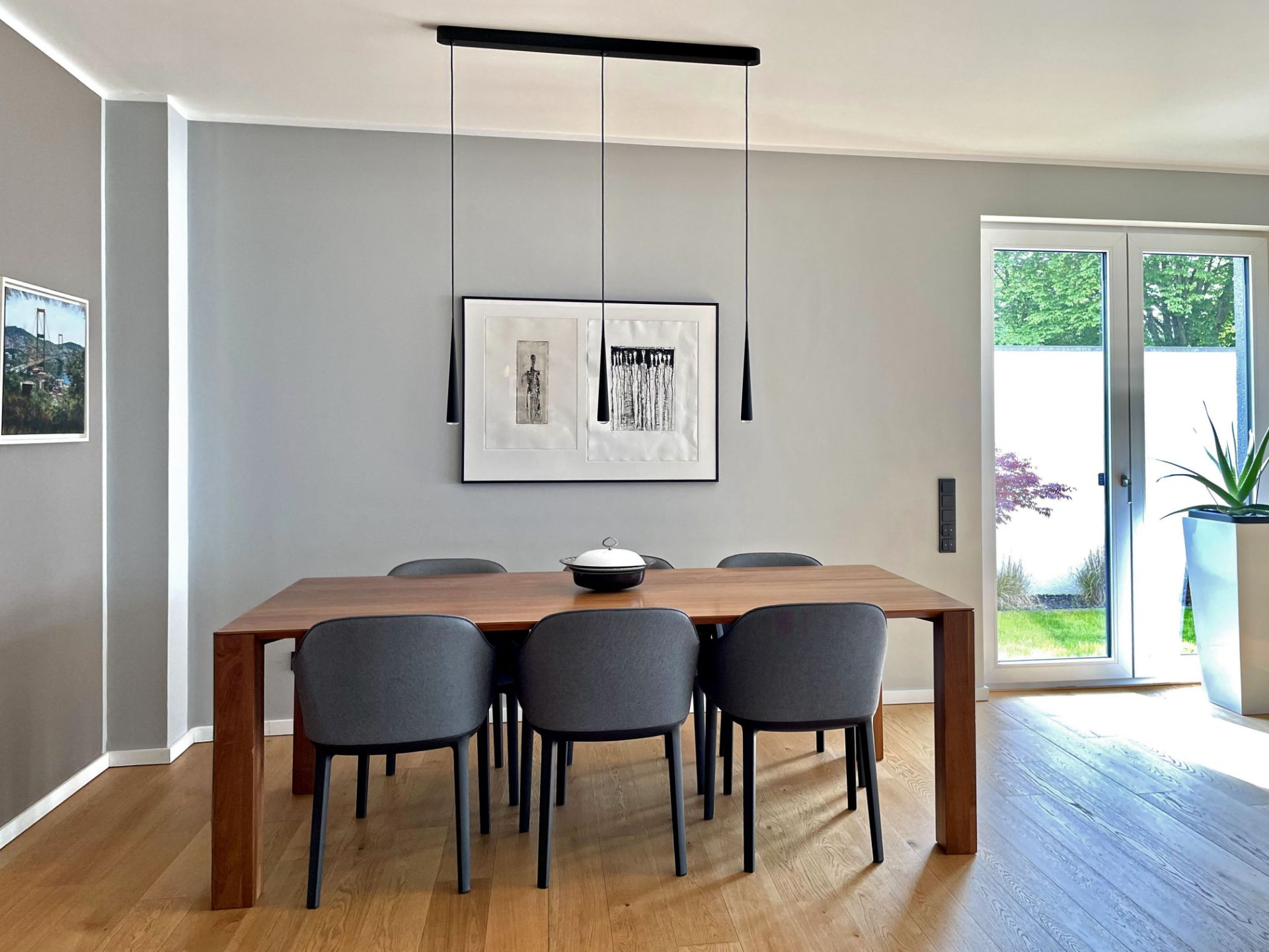 Esszimmer mit Holztisch, sechs grauen Stühlen, Wandkunst und großen Fenstern mit Pflanzen.