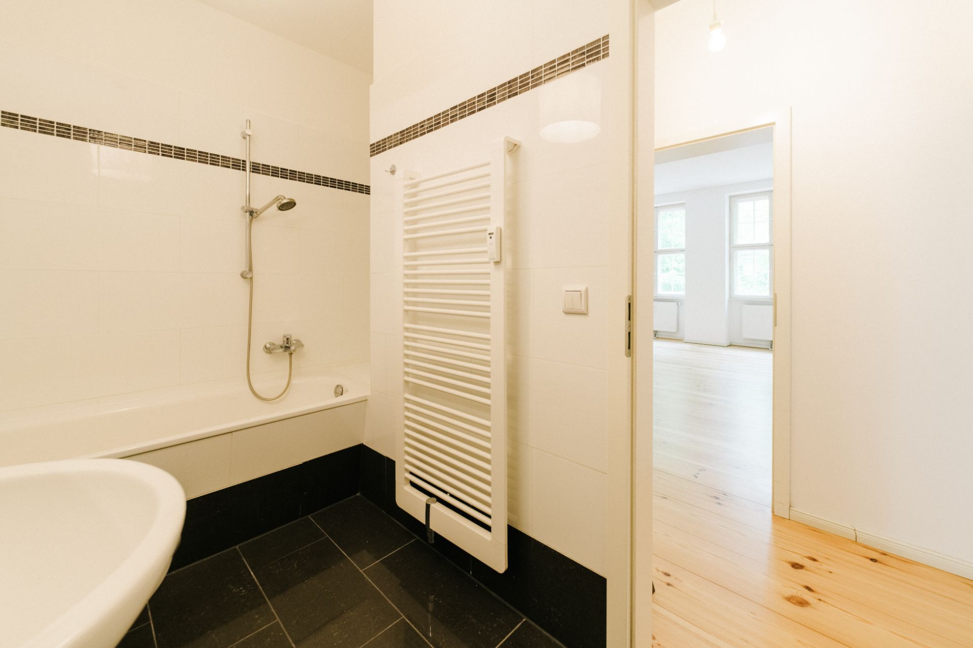 Helles Badezimmer mit Badewanne, Duschkopf und Handtuchheizkörper. Tür zum hellen Wohnbereich.