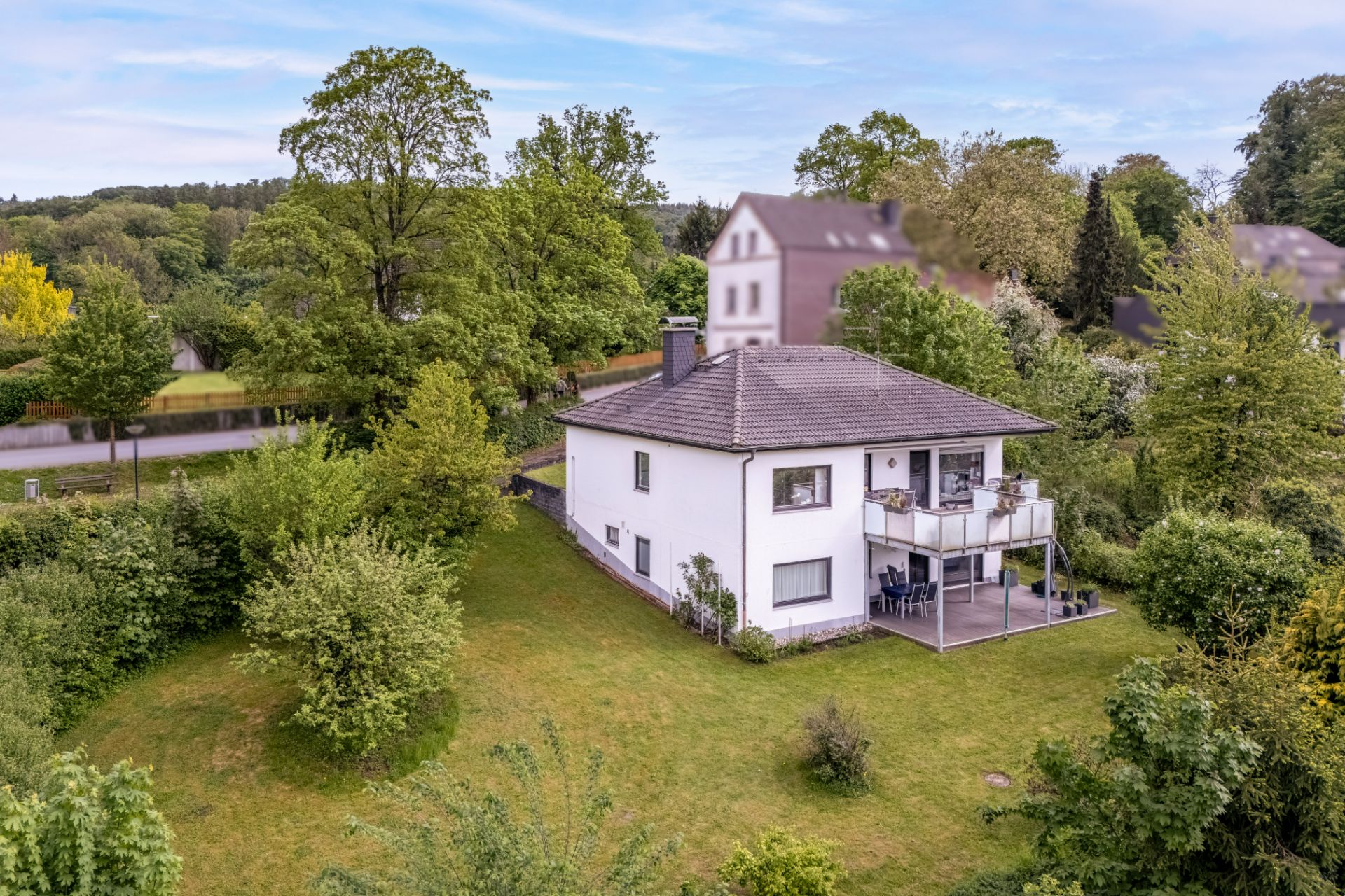 Zweistöckiges weißes Haus mit Balkon, umgeben von großem Garten und vielen Bäumen.