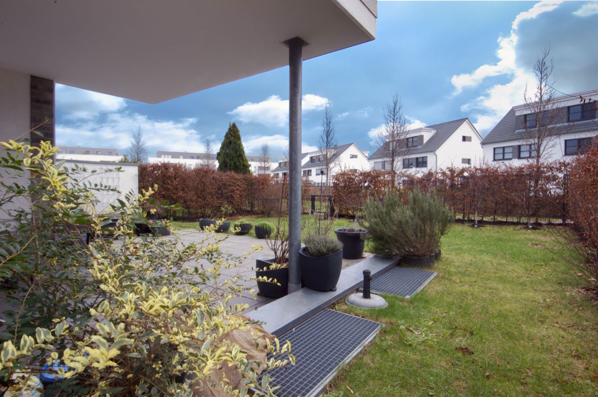 Blick auf eine Terrasse mit Pflanzen und angrenzendem Garten in einem Wohngebiet.